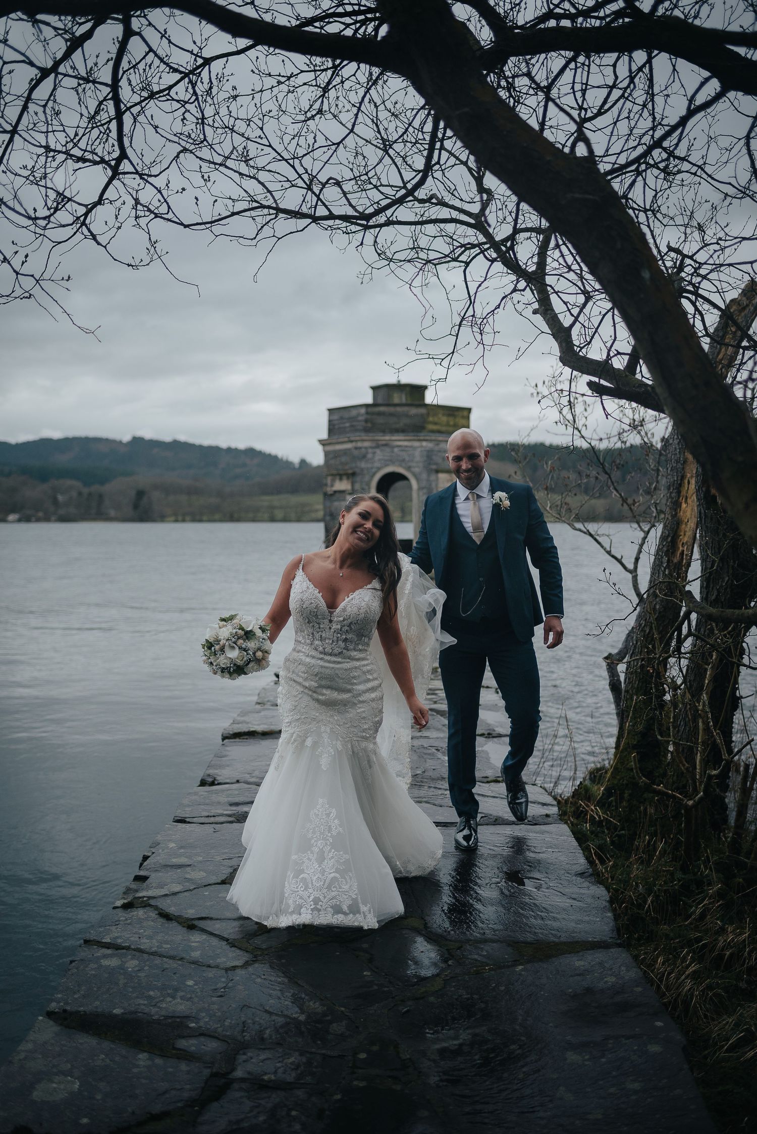 Newlyweds share playful moment on wet lakeside path under moody winter sky.
