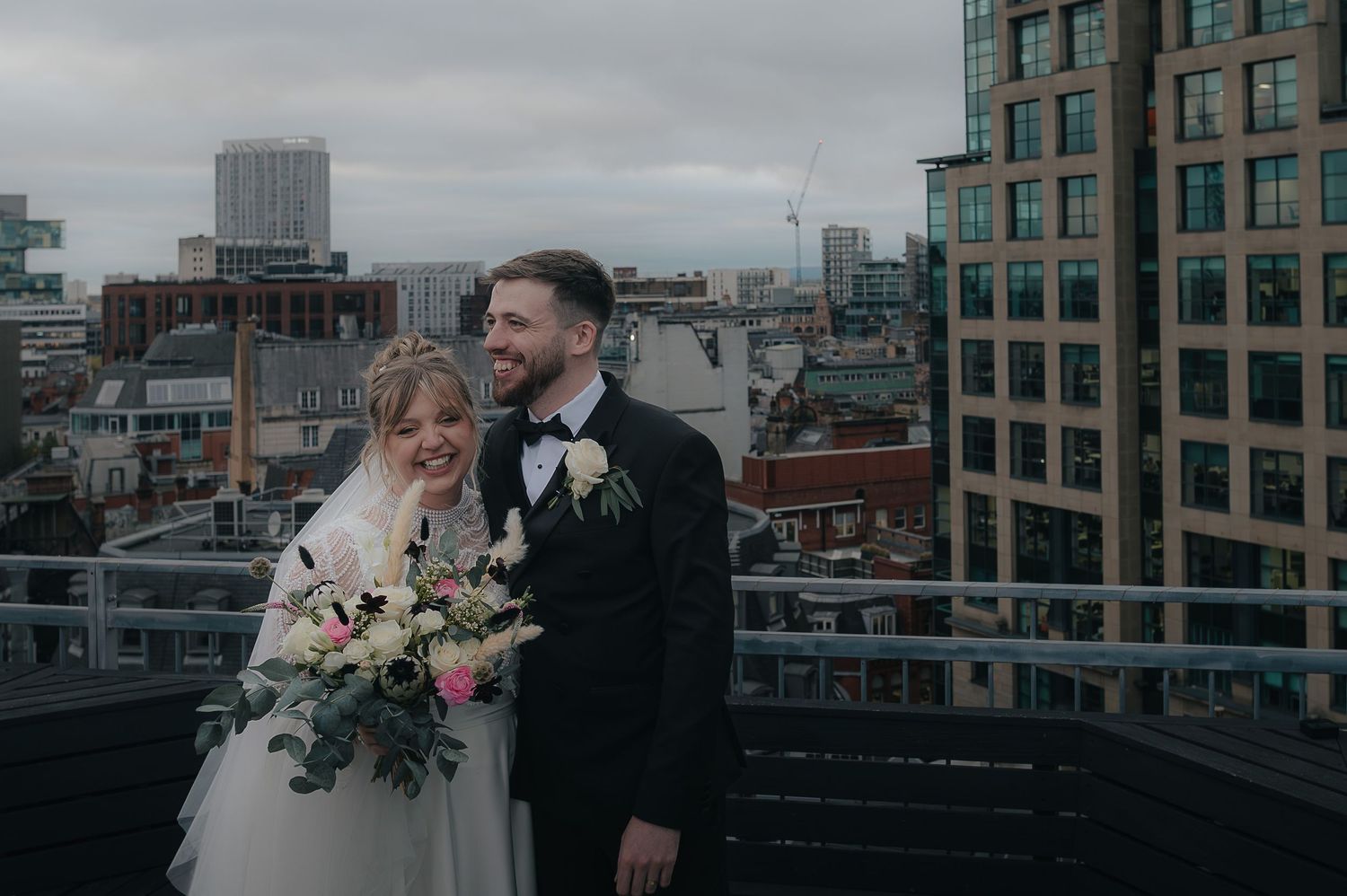 Couple embraces during romantic rooftop wedding ceremony with cityscape backdrop in overcast urban setting.