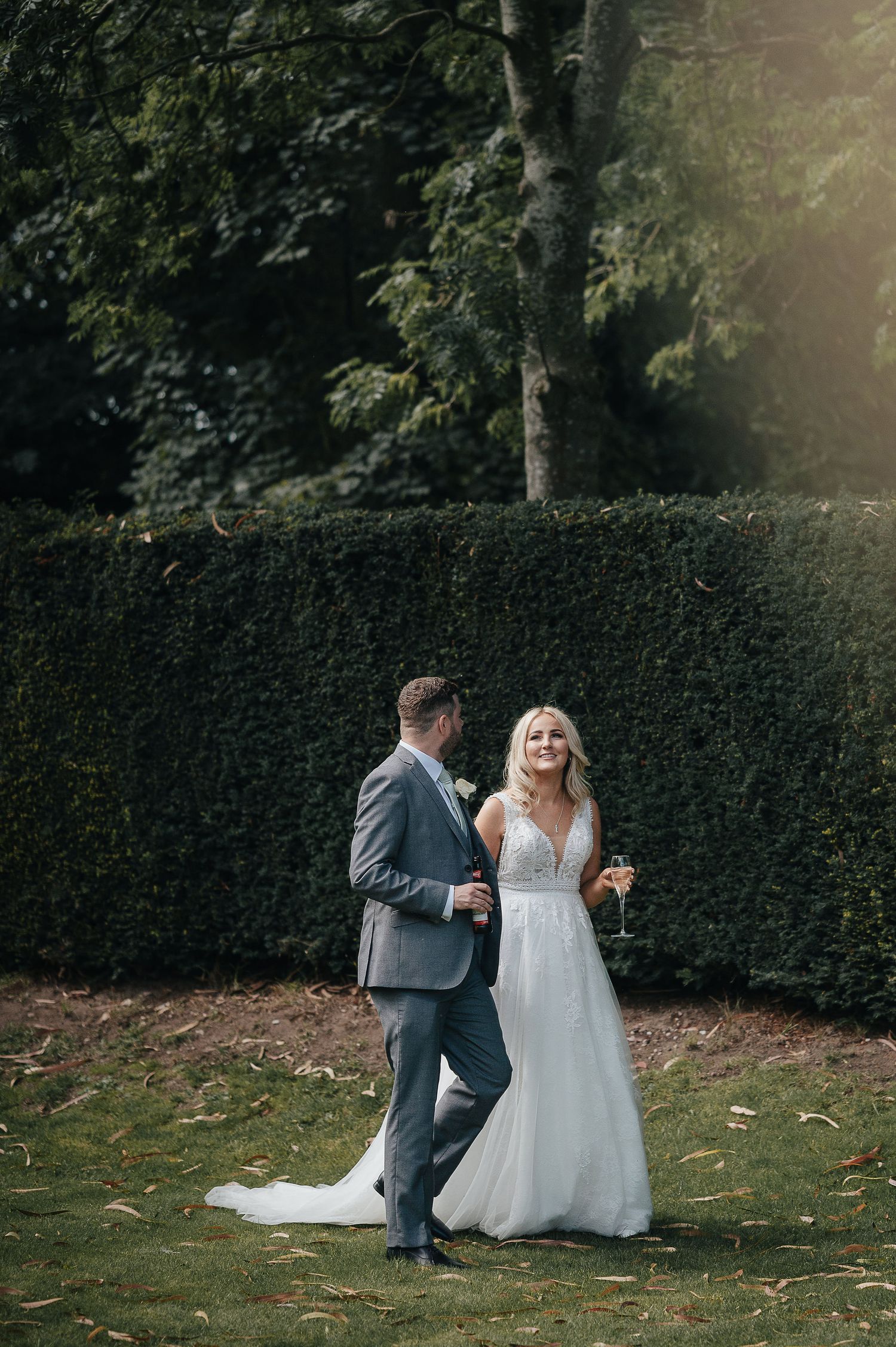 A happy wedding couple share a joyful moment on a lawn with a tall hedge background.