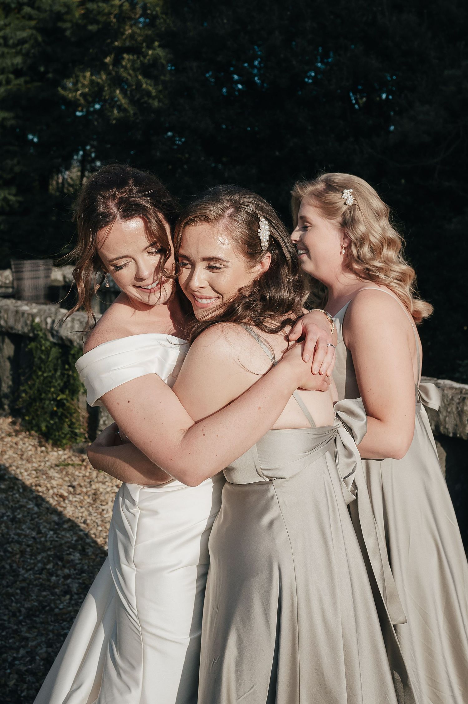 Three bridesmaids in light colored dresses share a joyful embrace in sunlit garden setting.