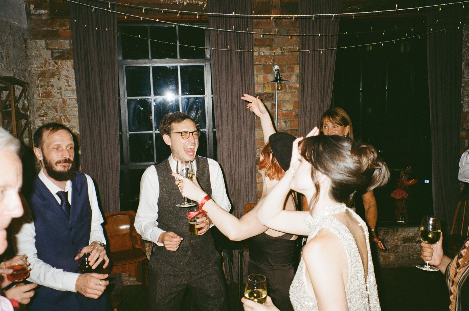 Guests dancing and celebrating at an indoor wedding reception with string lights and rustic brick walls.