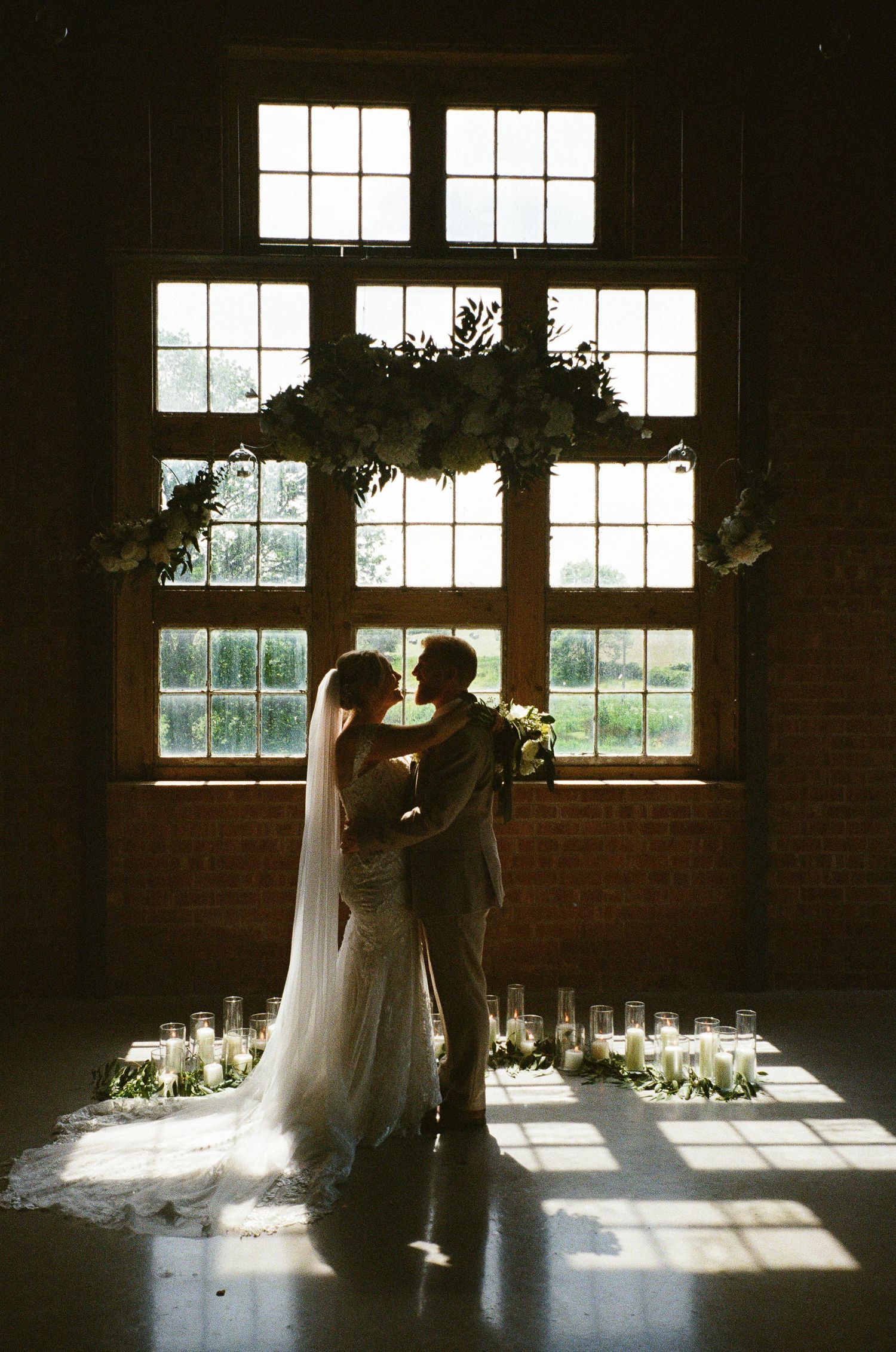 Bride and groom silhouetted kissing before large industrial windows with floral arch and candles at elegant wedding ceremony.