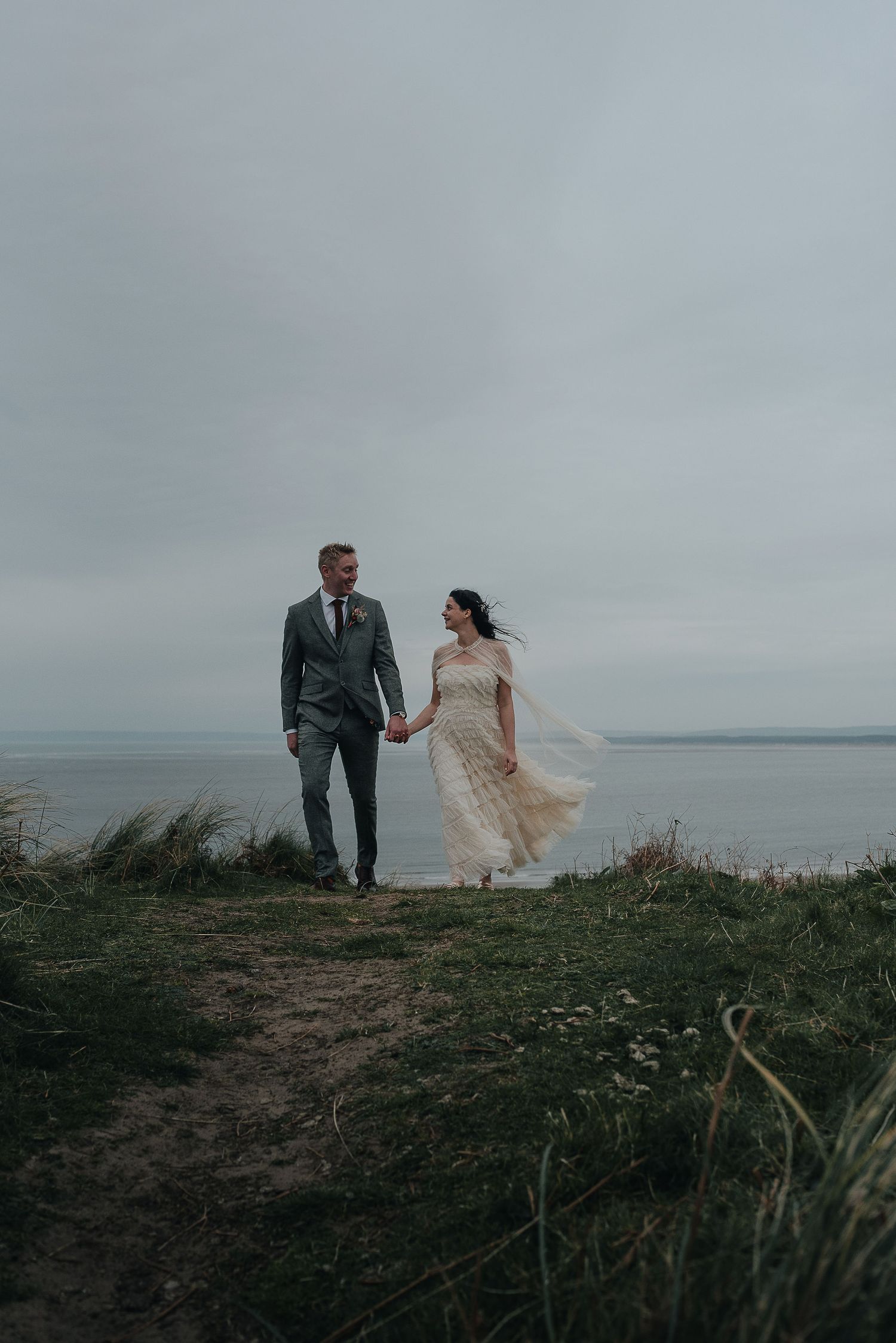 A couple holds hands on a coastal path as the bride's dress flows in the wind.