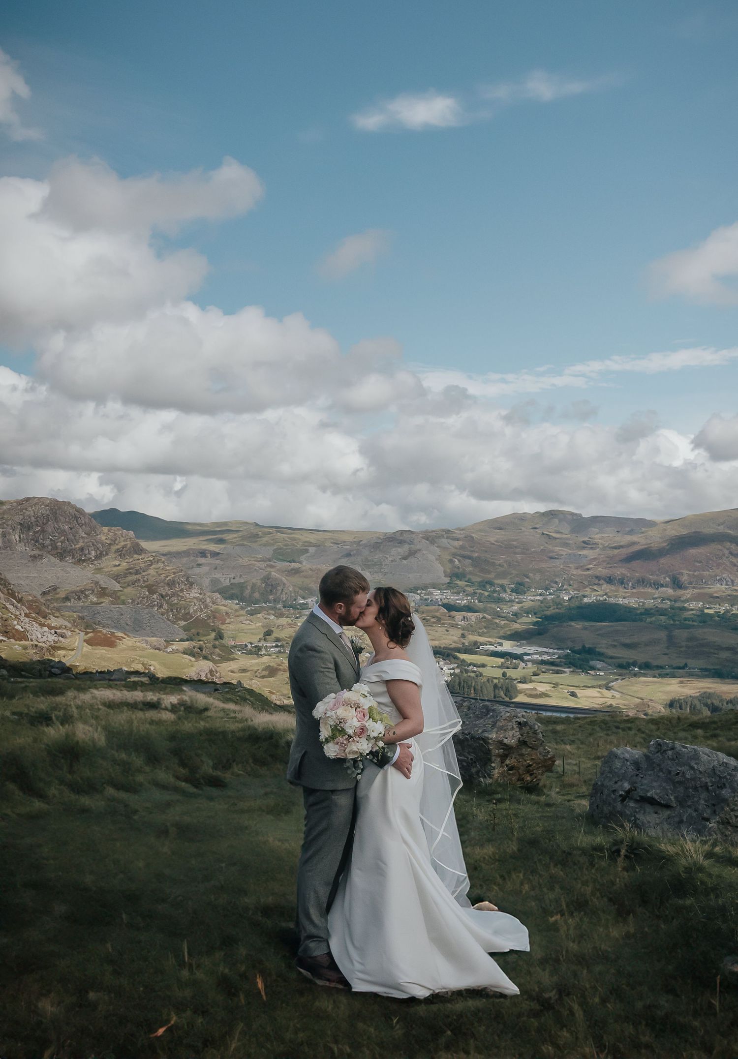A wedding couple stands on a hillside with dramatic mountain landscapes and clouds in the background.