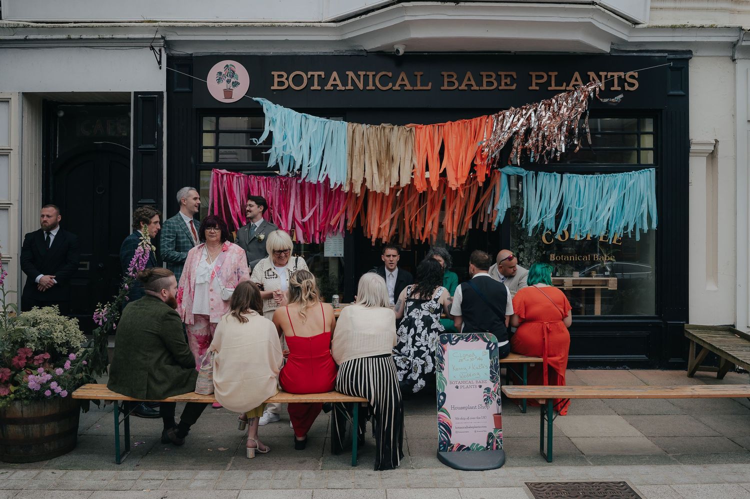 Wedding guests seated outside a botanical shop with colorful hanging decorations.