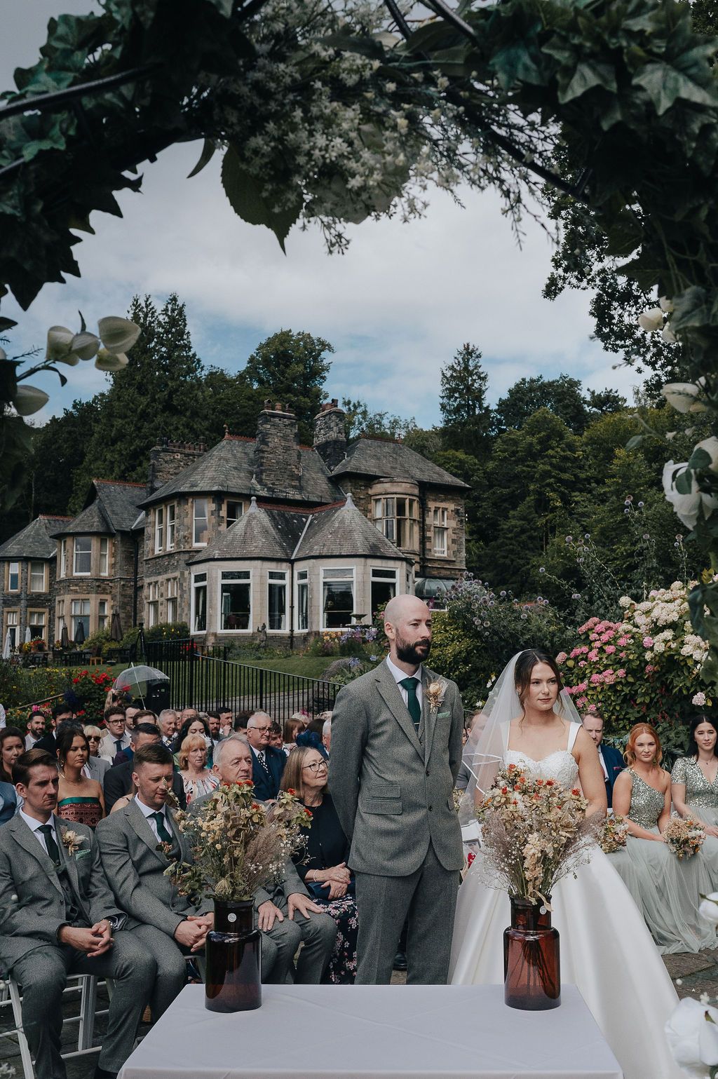 Outdoor wedding ceremony in front of a grand stone mansion with floral decorations.