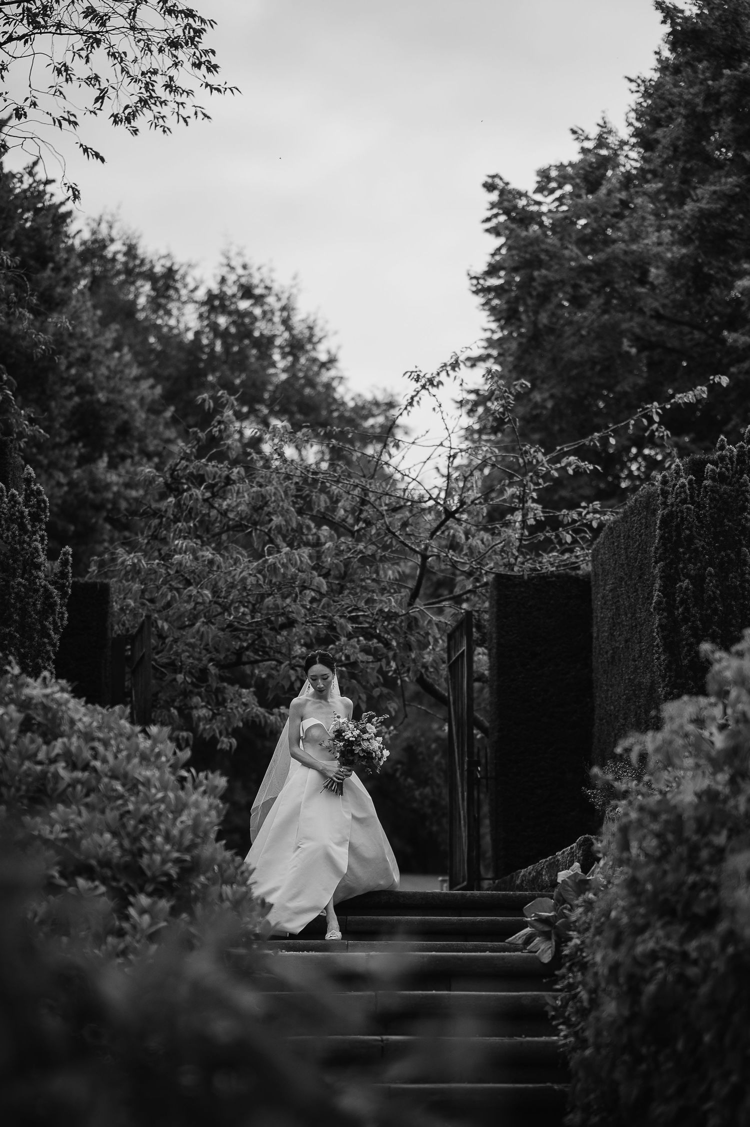 Black and white photo of a bride ascending stone garden steps surrounded by lush foliage.