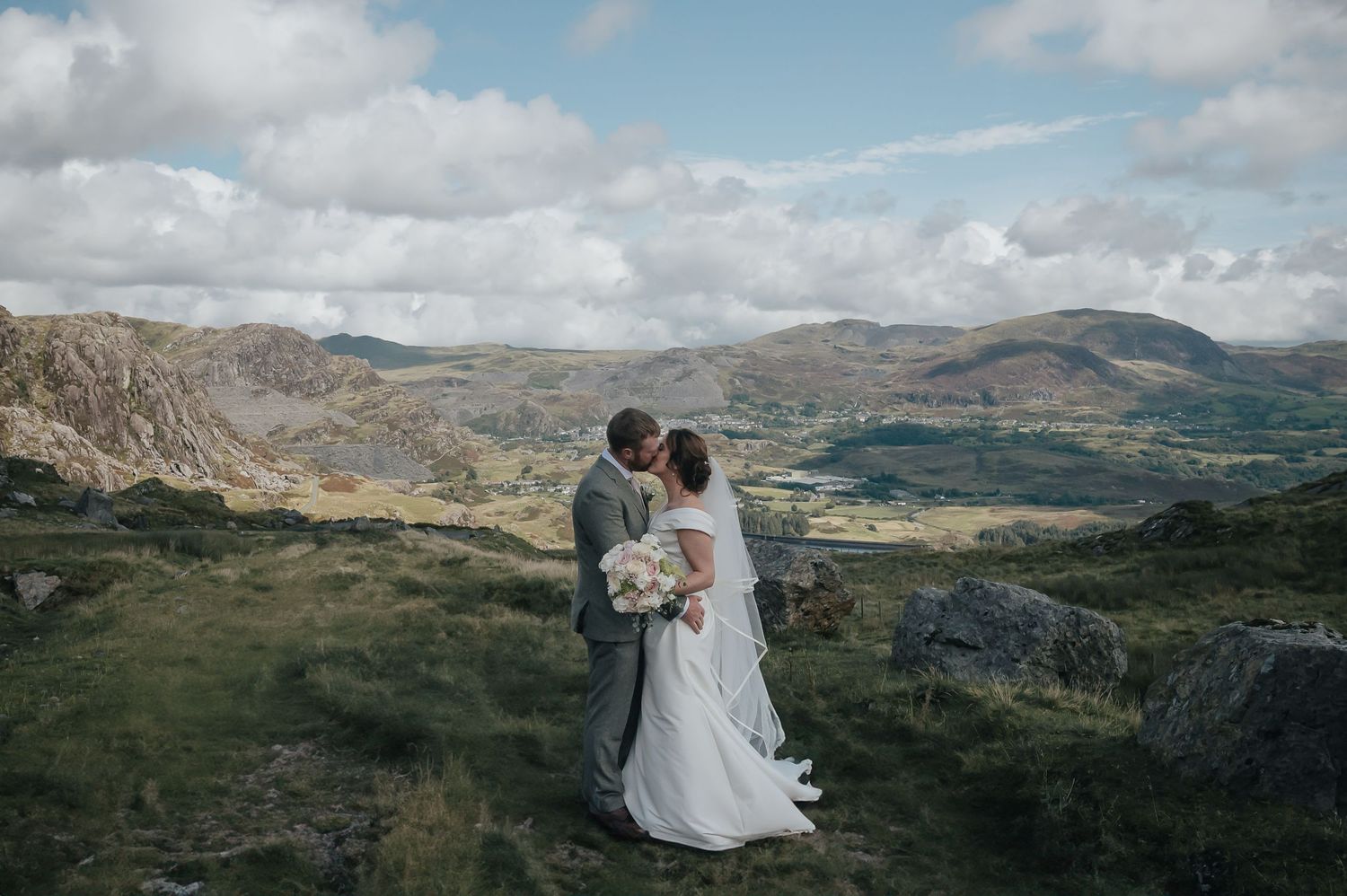Couple in wedding attire embrace while overlooking dramatic mountain landscape.
