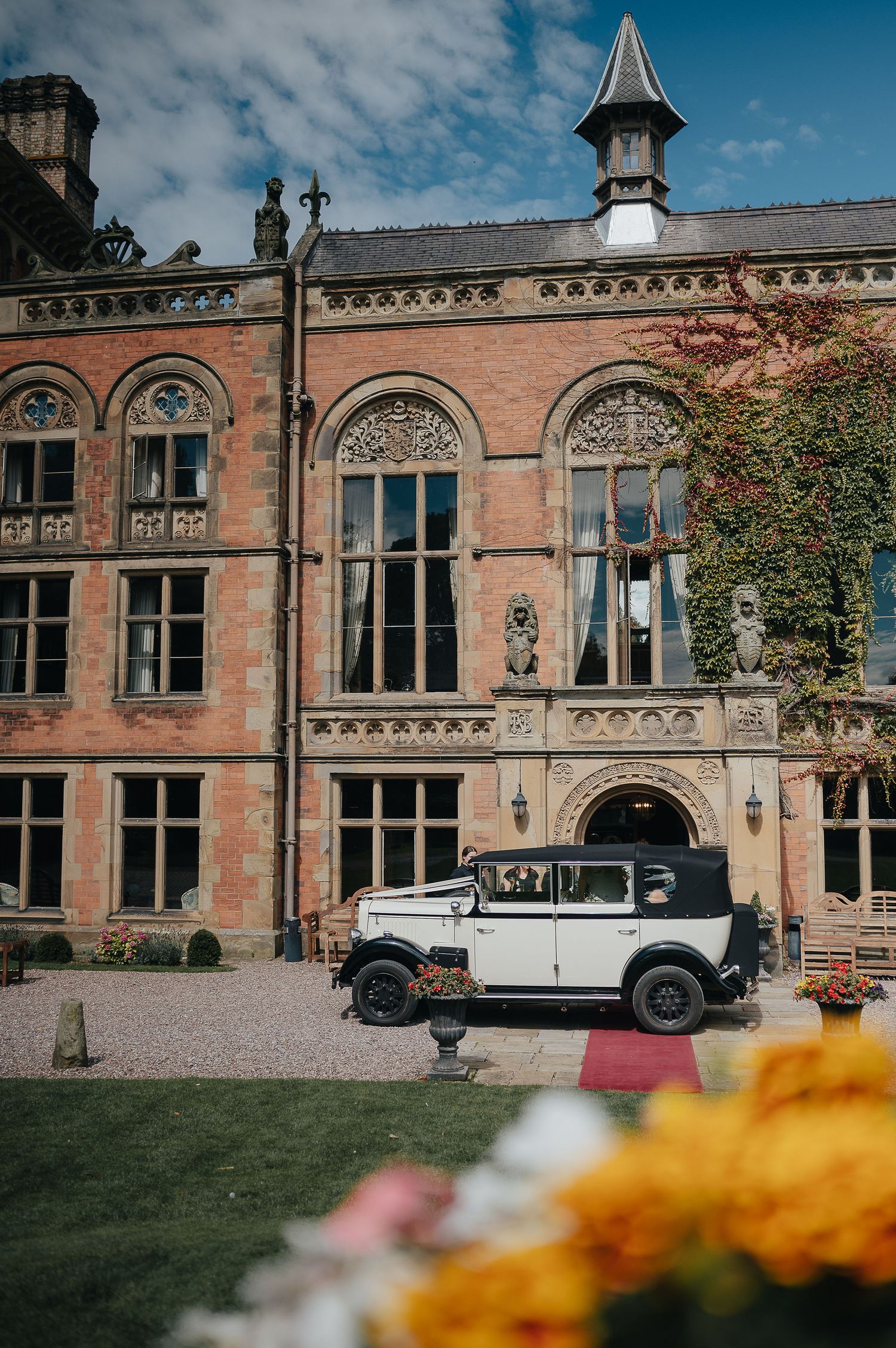 Classic white vintage car parked in front of a grand Gothic revival brick mansion.