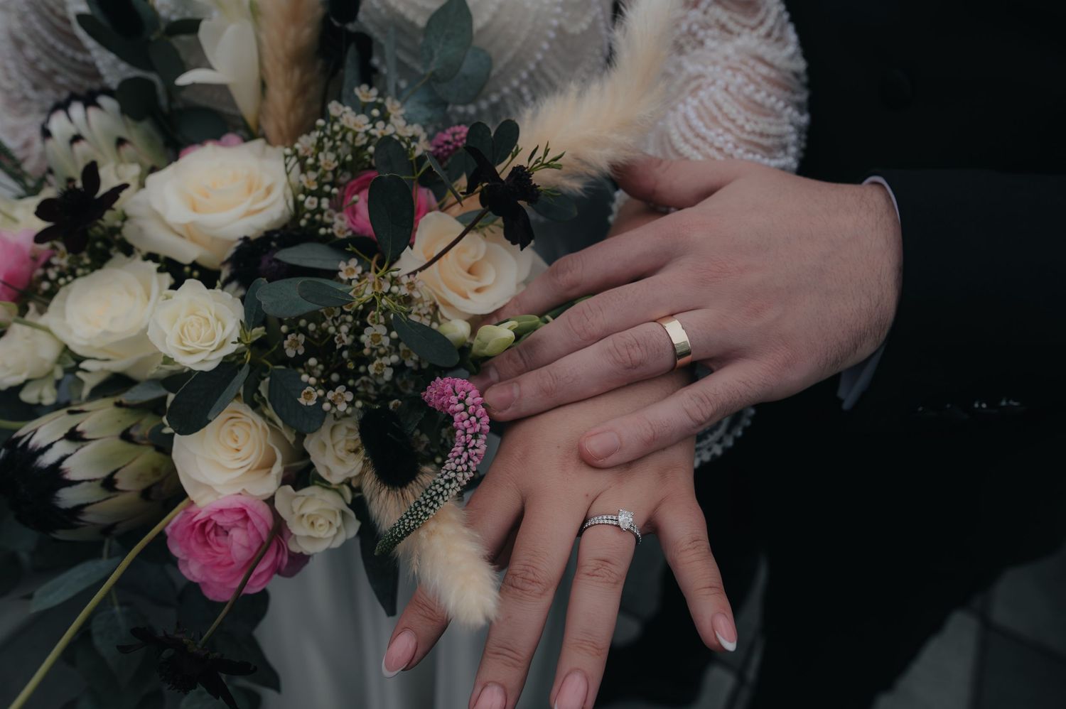 Hands wearing wedding rings displayed over elegant bridal bouquet with white and pink roses and greenery.