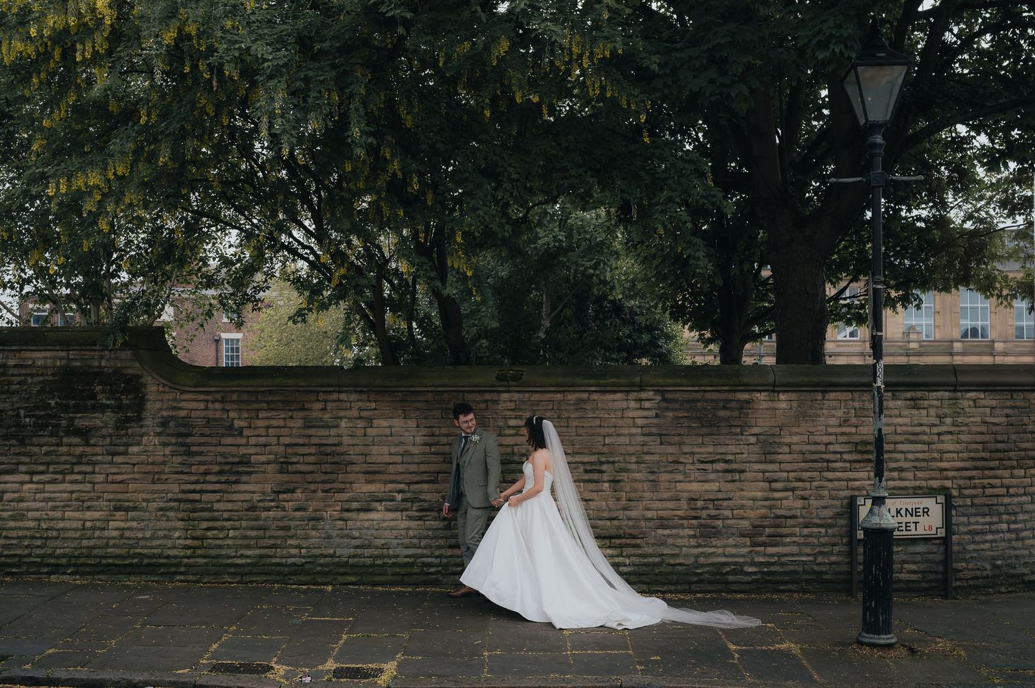 A elegant white wedding gown flows across a brick-lined street against a rustic wall backdrop.