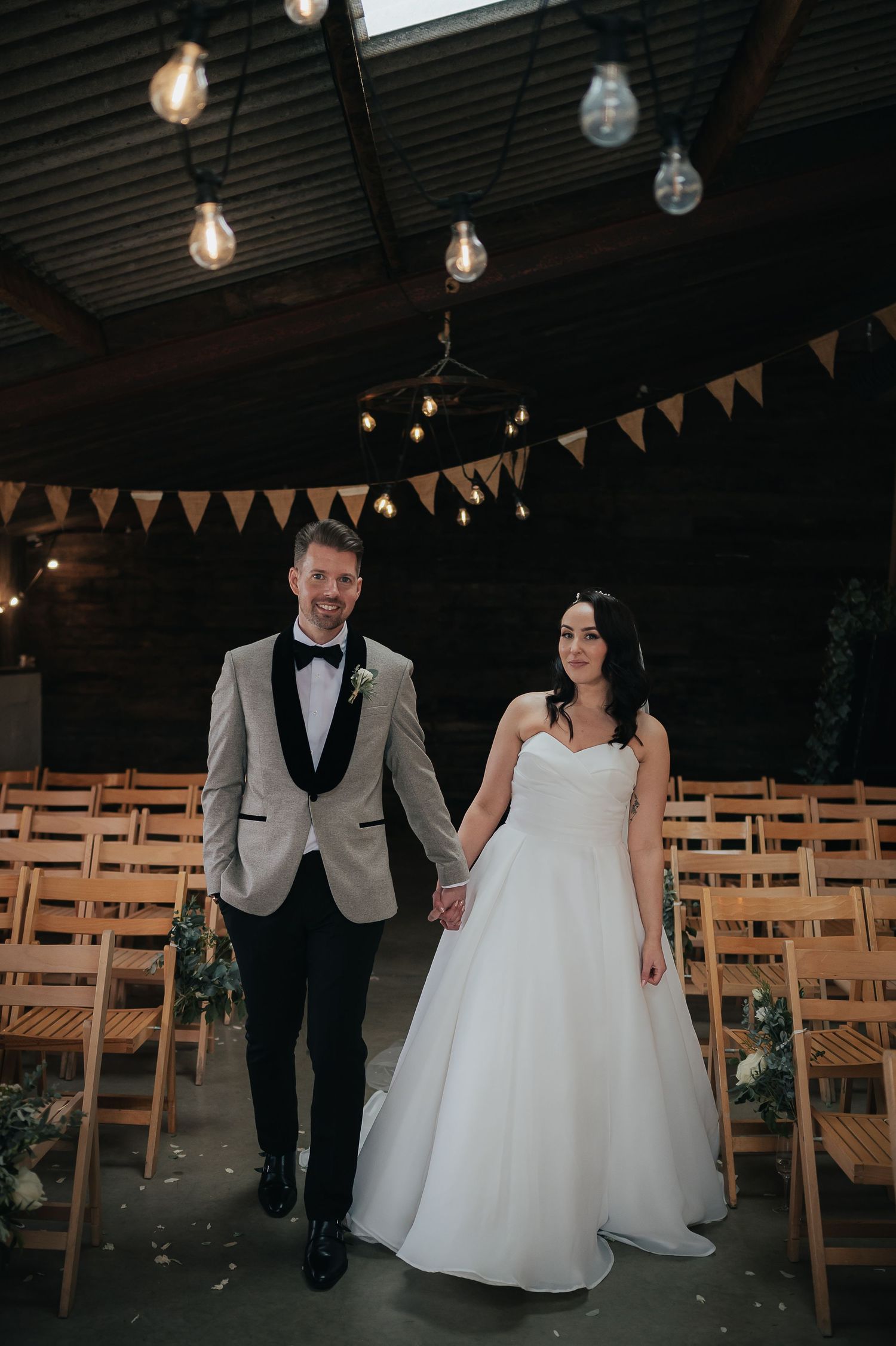 Couple walks through rustic indoor venue with string lights and bunting decorations.