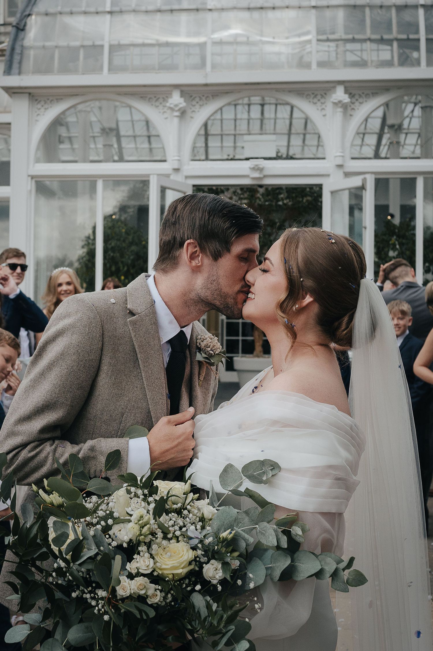 A tender wedding moment captured in front of an elegant conservatory with white arched windows.
