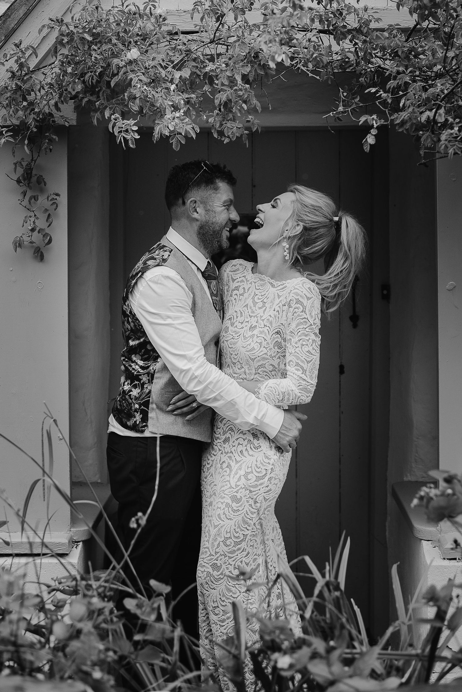 Happy wedding couple laughing together under an ivy-covered archway in black and white.