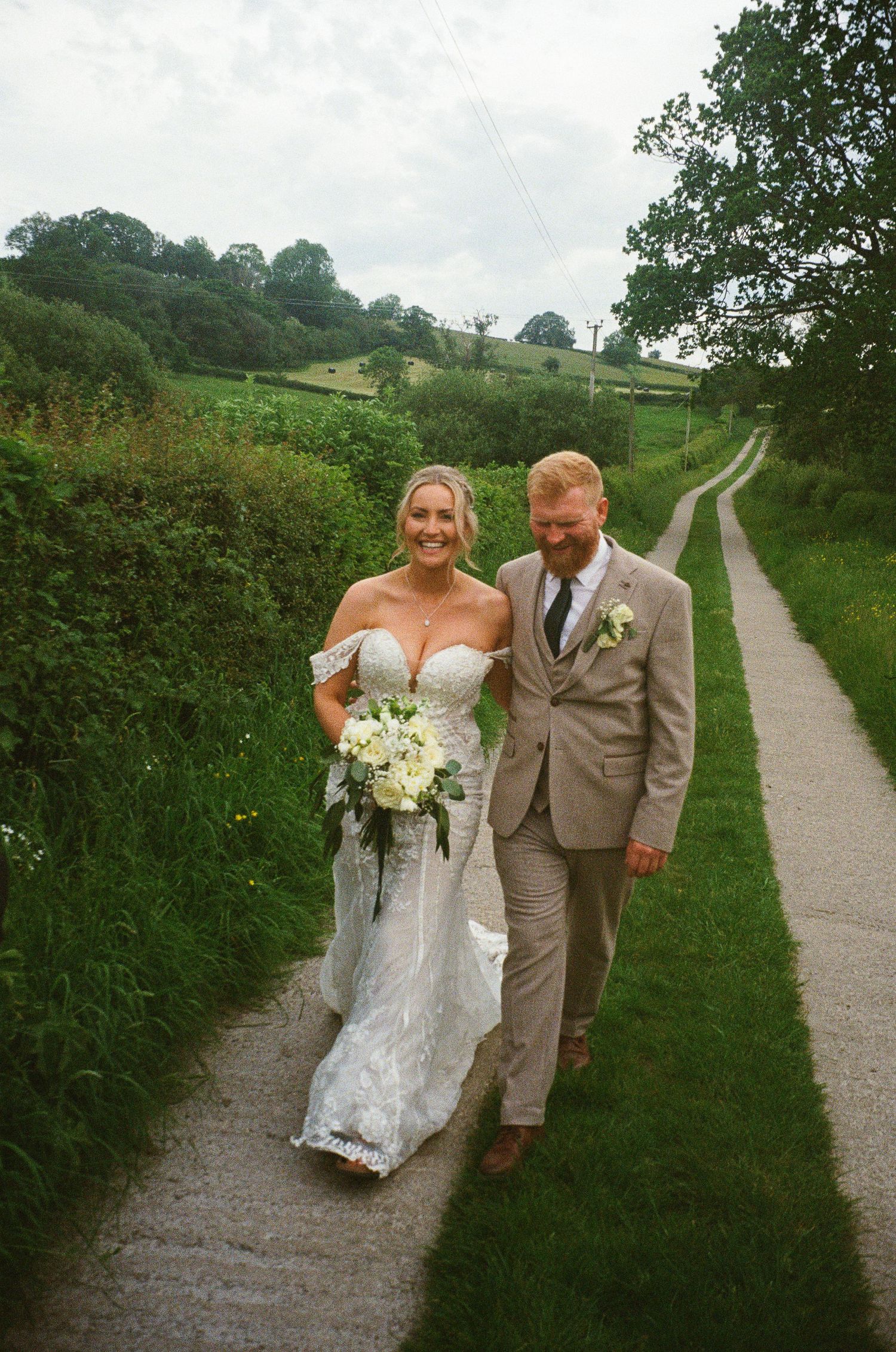 Bride in white lace gown holding bouquet walks with groom in tan suit along a scenic countryside path.