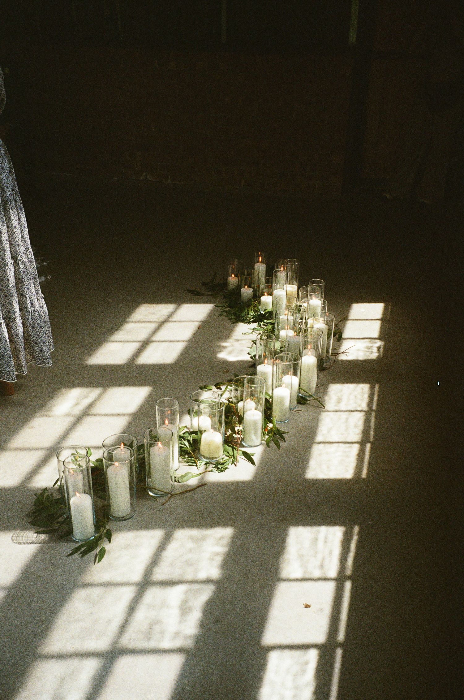 White pillar candles in glass holders arranged on floor with greenery, casting shadows in warm sunlight at elegant wedding ceremony.