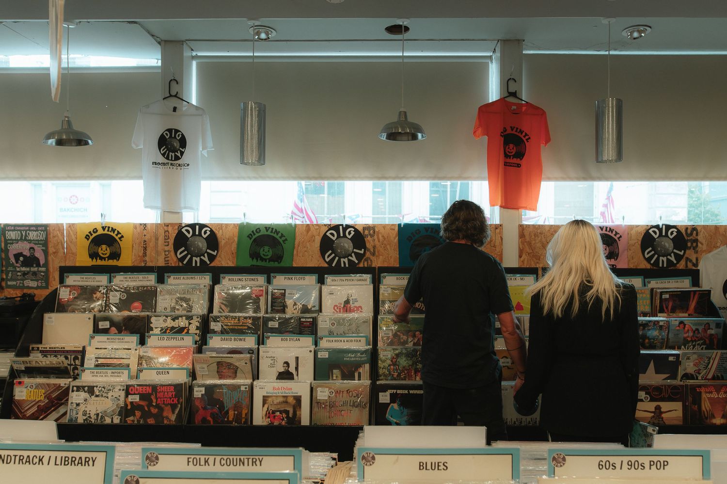Two people browse vinyl records in a music store with colorful album covers displayed on shelves under overhead lighting.