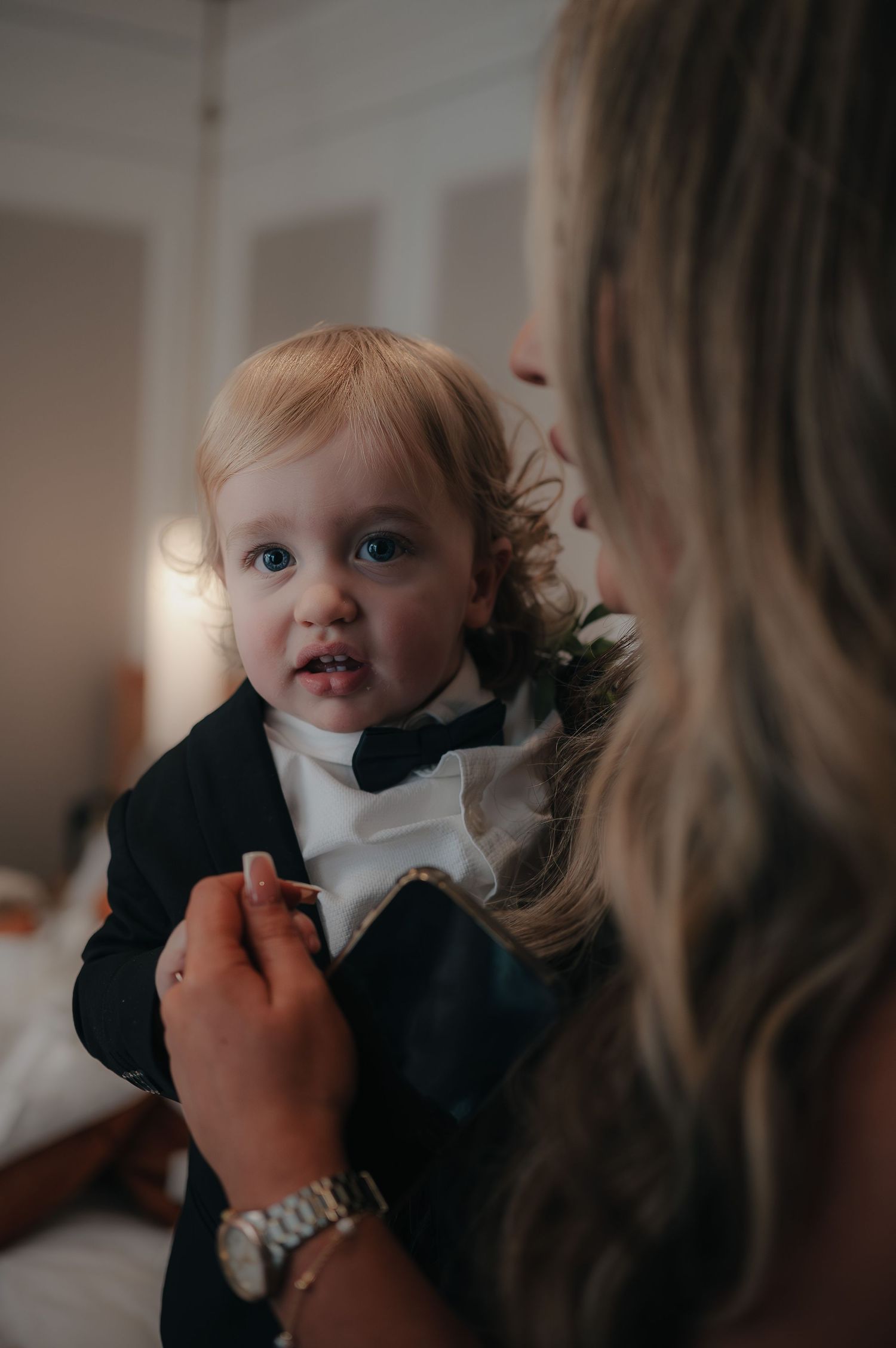 An adorable baby in a formal black and white suit with bowtie being tended to, lit with soft natural lighting.