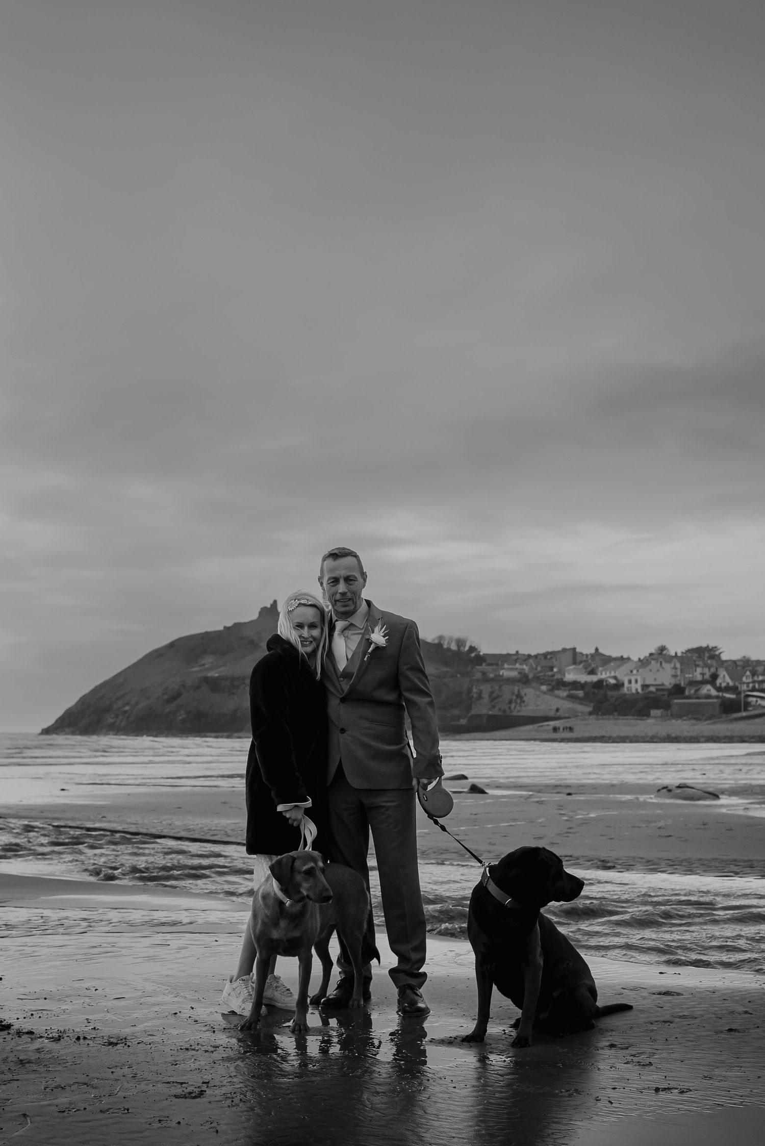 Couple poses with their two dogs on beach with coastal headland in background.