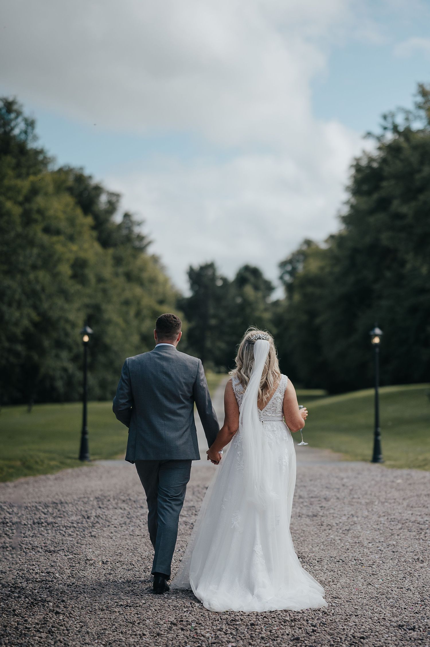 A newlywed couple walks hand in hand down a tree-lined gravel path.