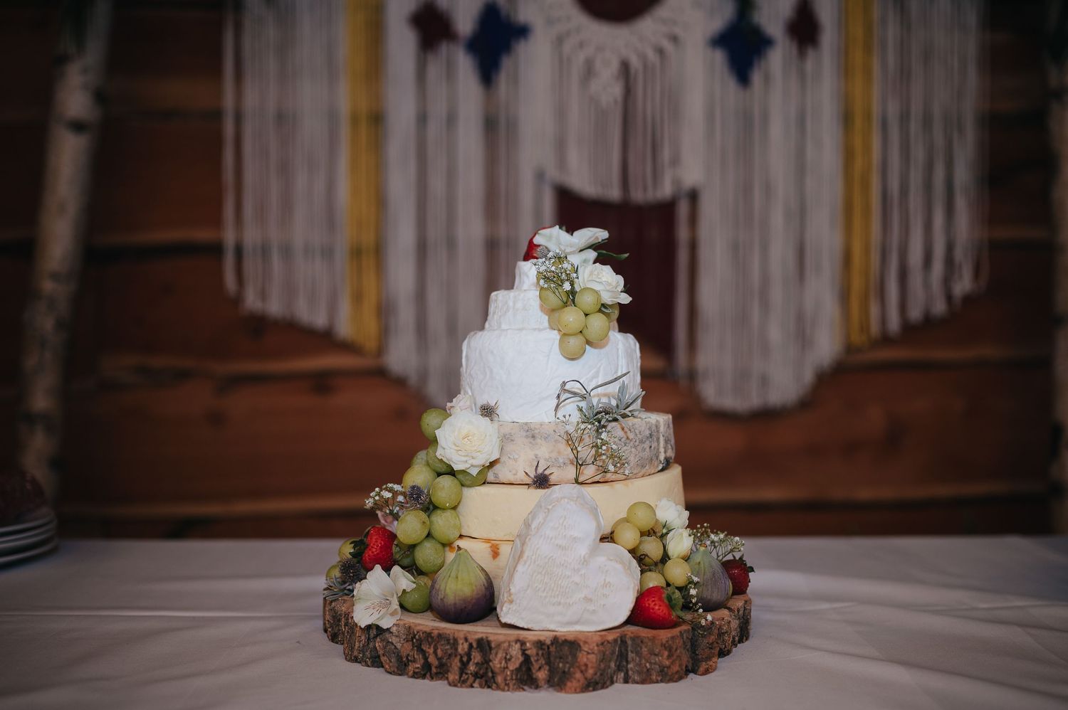 A rustic wedding cake displayed on a wooden slice with fresh fruit and cheese wheels arranged in tiers.