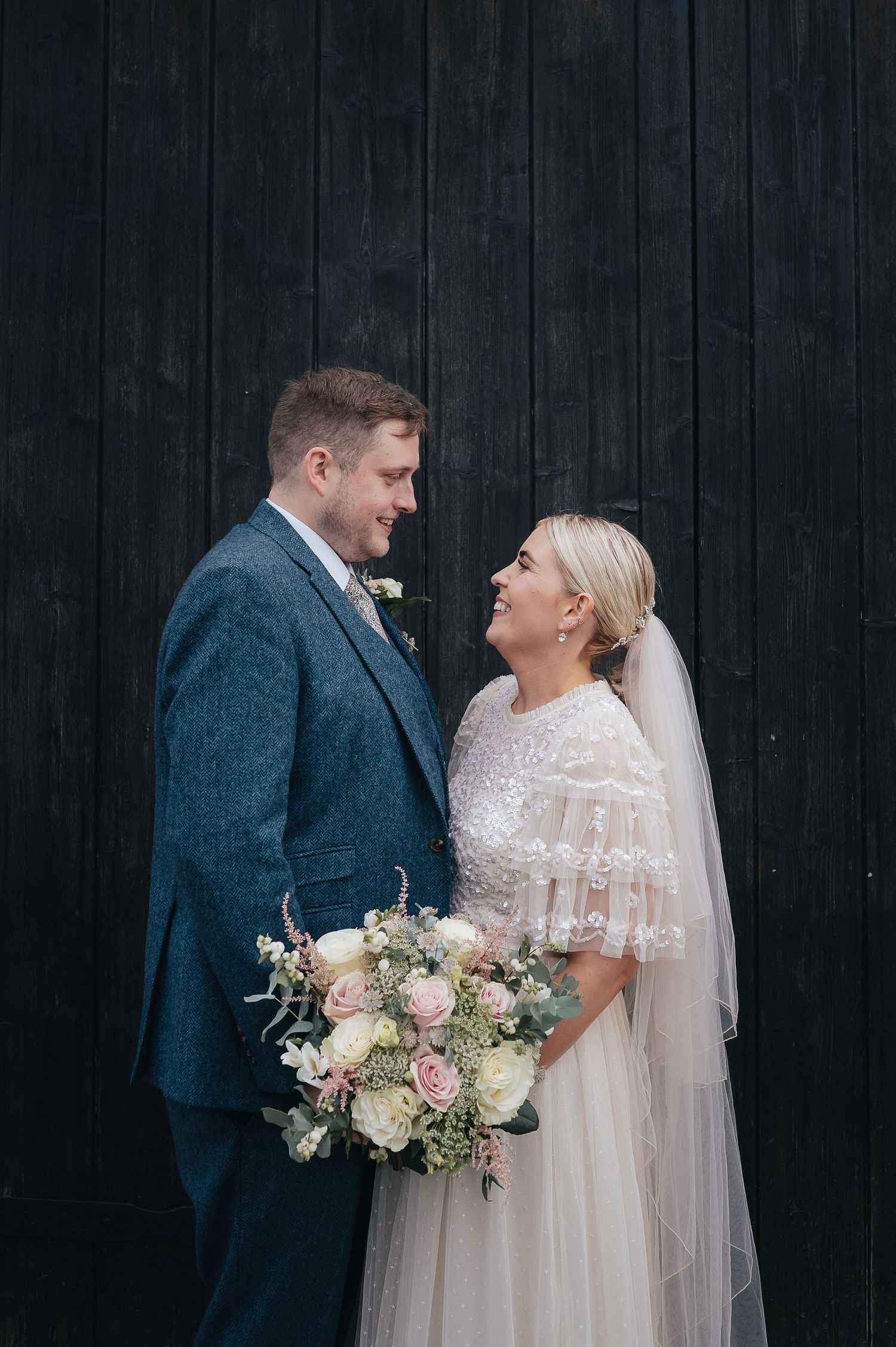 A wedding couple shares a joyful moment against a dark wooden wall, with a beautiful pastel floral bouquet.