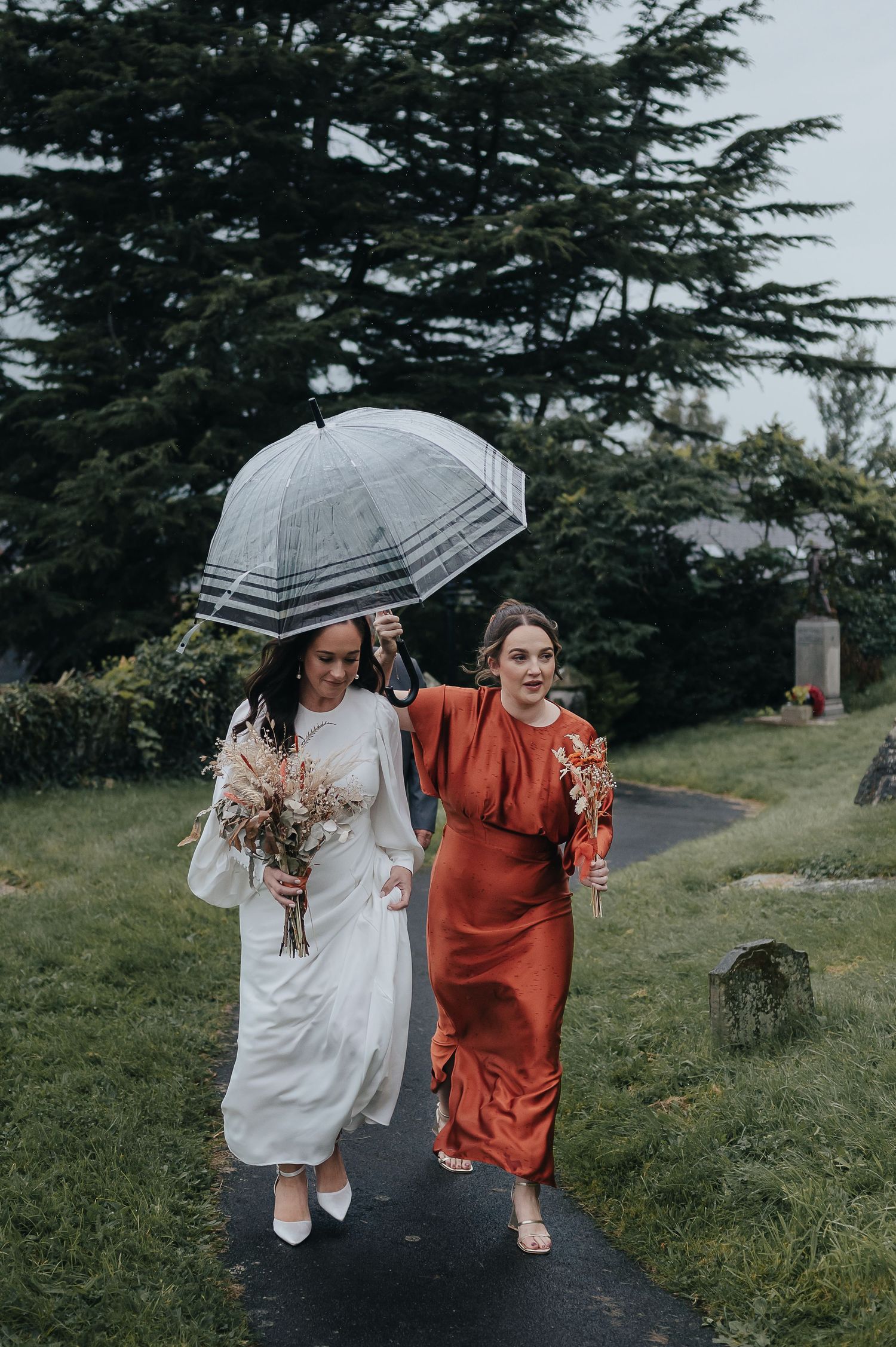 Two wedding guests walk along a path sharing a clear umbrella in the rain.