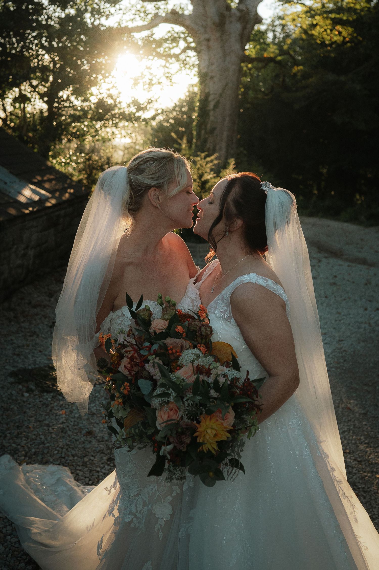 Two brides share a romantic moment at sunset while holding a vibrant fall-colored bouquet in their wedding gowns and veils.