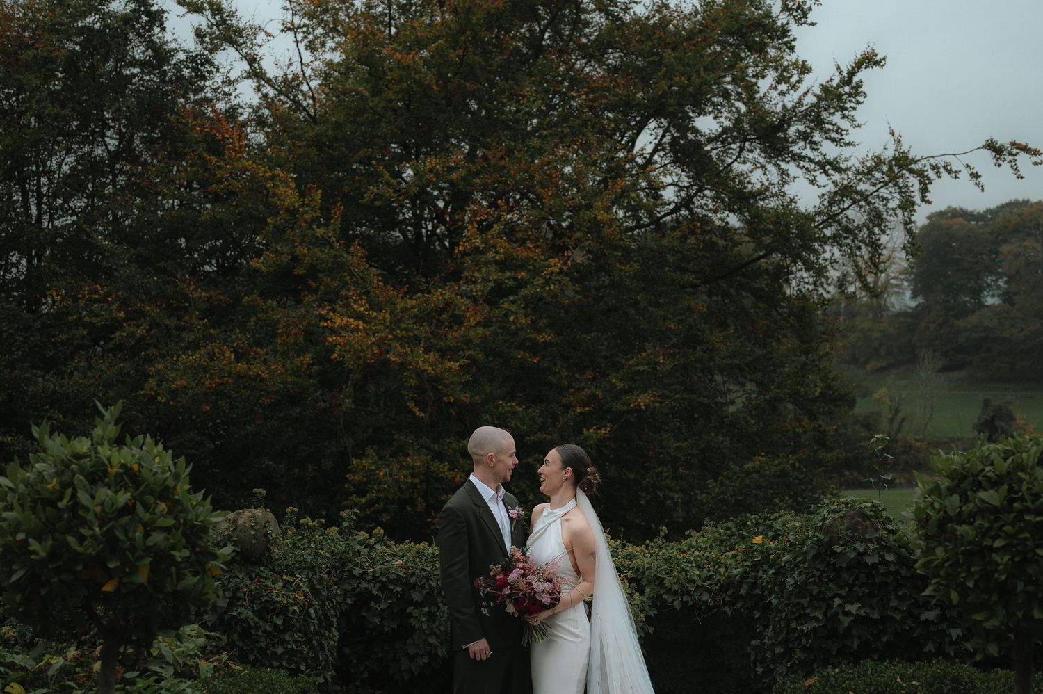 A romantic wedding couple embraces outdoors surrounded by lush autumn trees and natural scenery.