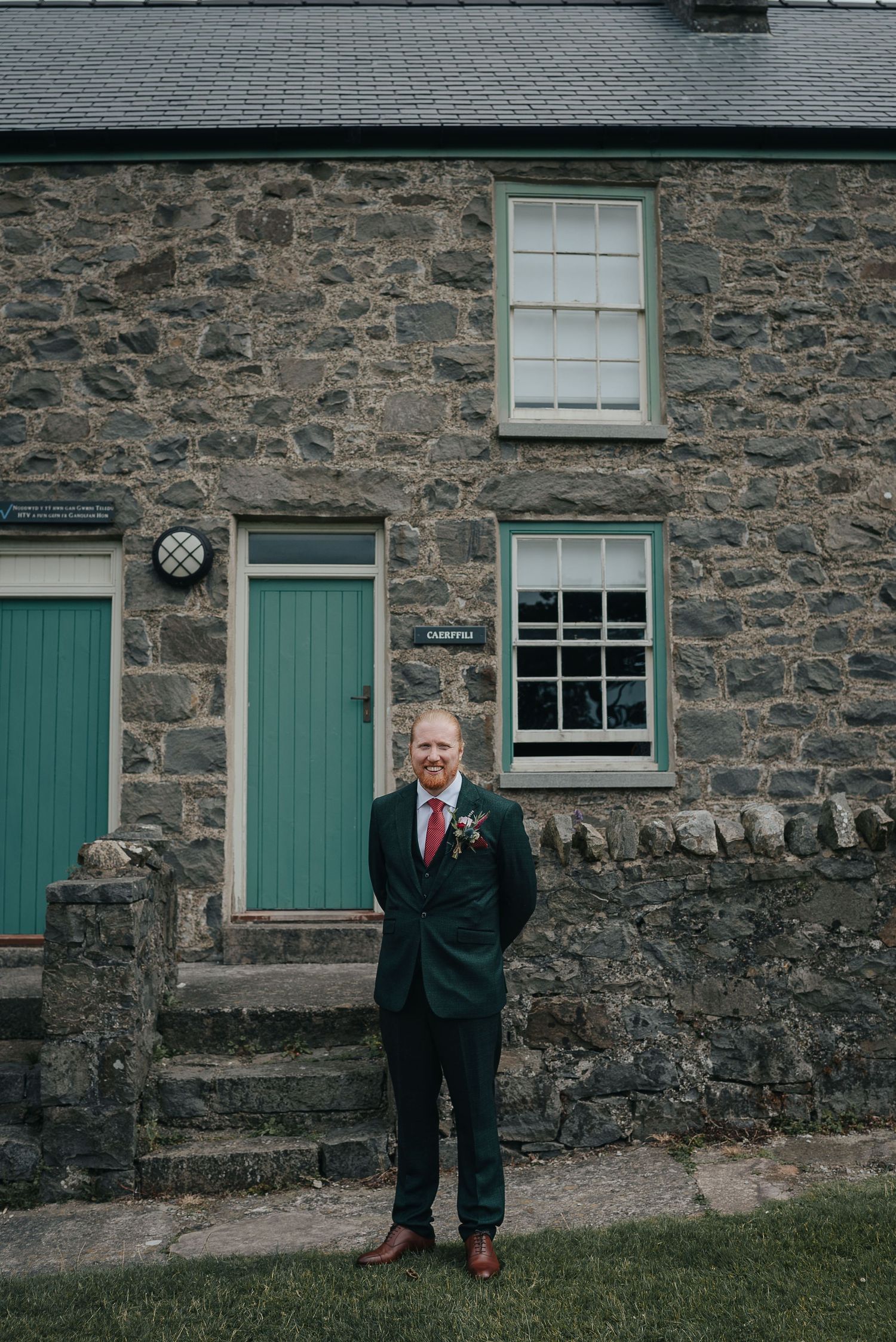 A person in formal attire stands in front of a traditional stone cottage with green trim.