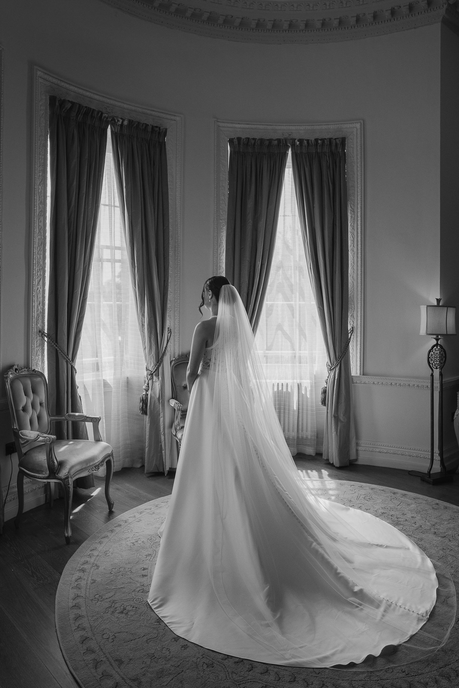 Black and white photo of wedding dress silhouette by elegant bay windows in classic room.