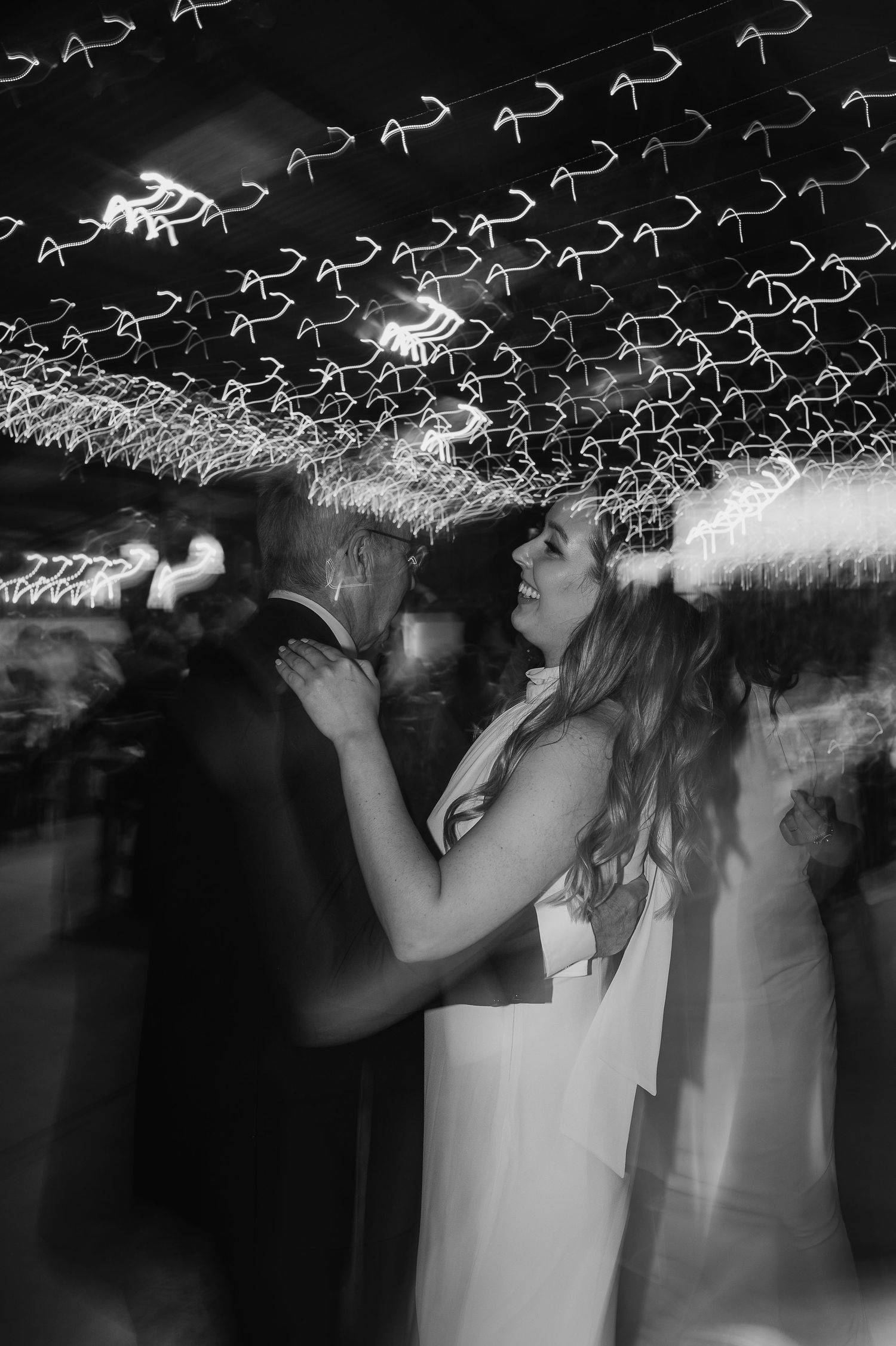 Black and white photo of a couple dancing beneath decorative string lights creating a dreamy pattern.