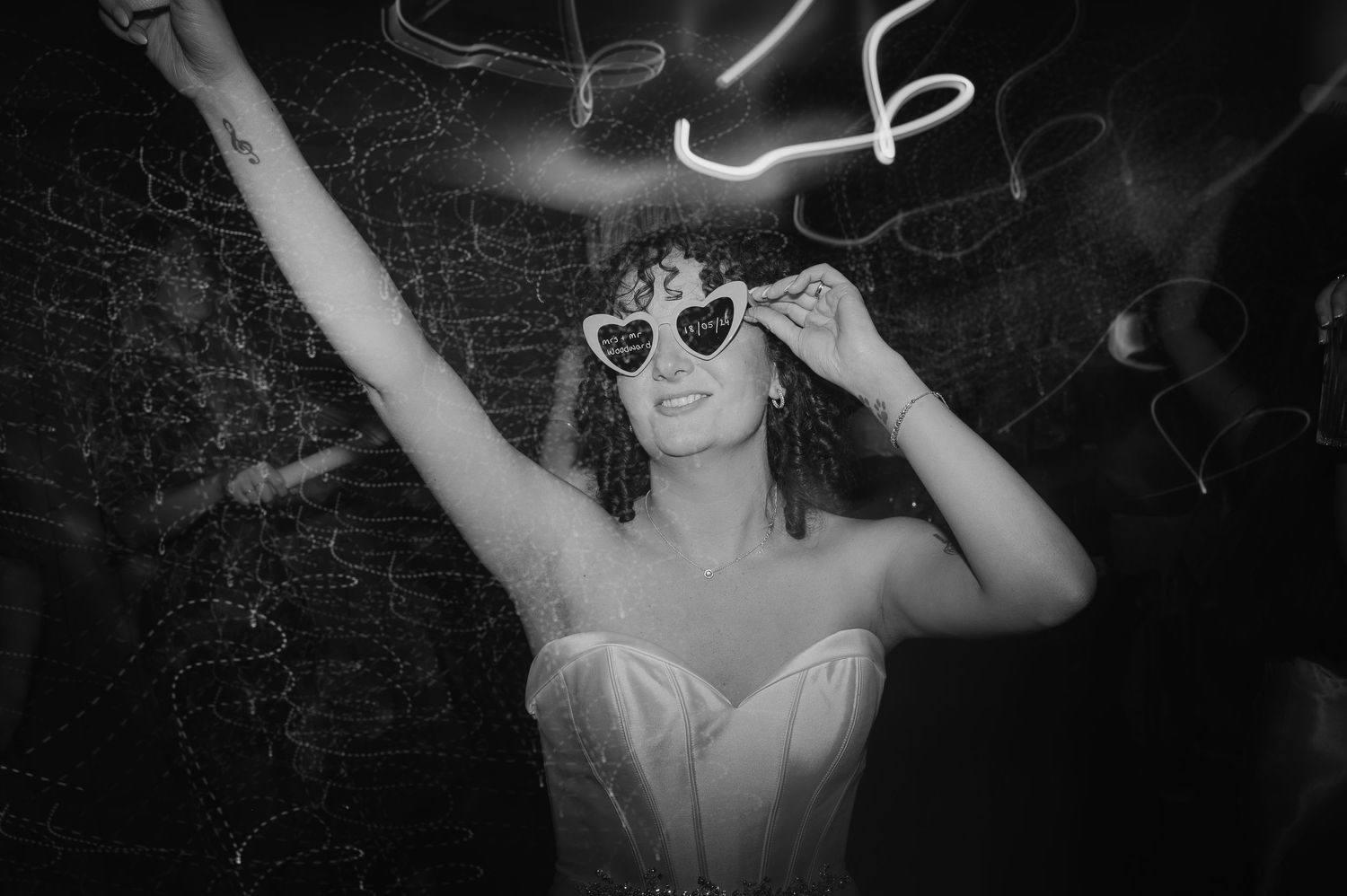 Person in a wedding dress wearing sunglasses poses with raised arms under neon lights.