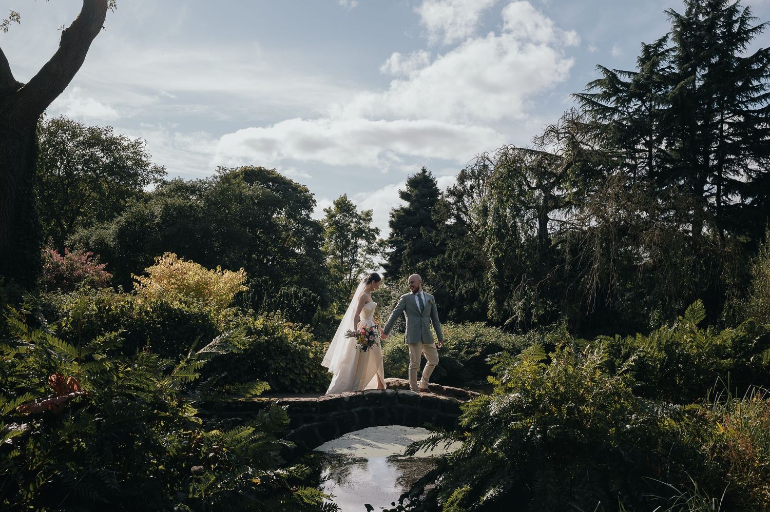 Two figures cross a small garden bridge in a lush green landscape.
