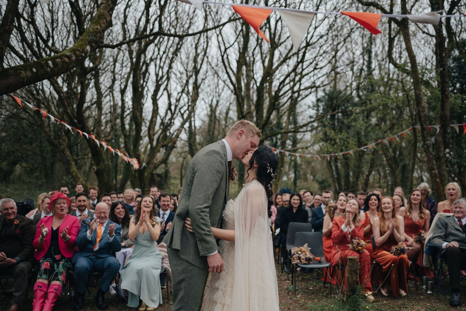 A wedding ceremony in a wooded setting with festive bunting decorations and gathered guests watching the couple.