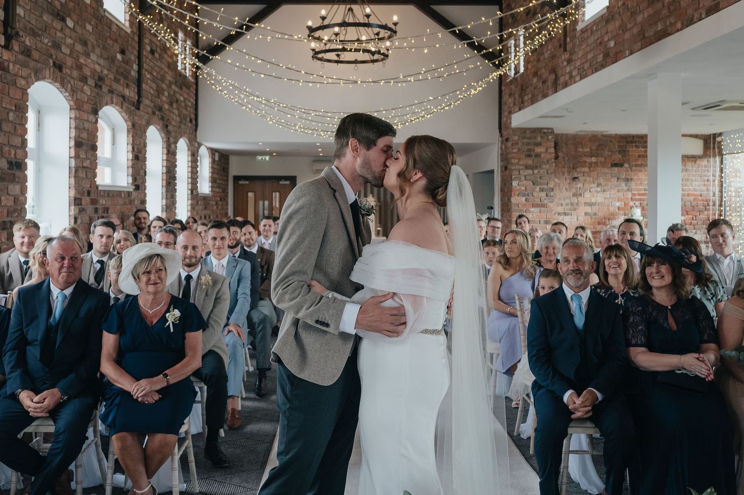 A couple shares their first kiss during an indoor wedding ceremony surrounded by guests in a rustic brick venue.