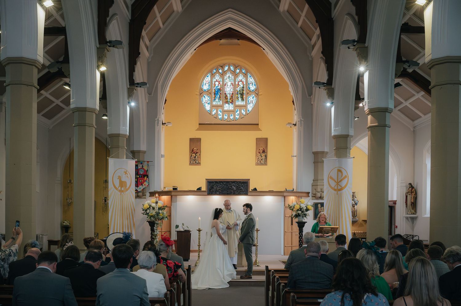 A wedding ceremony taking place in a beautiful church with Gothic arches and a yellow-tinted altar area.