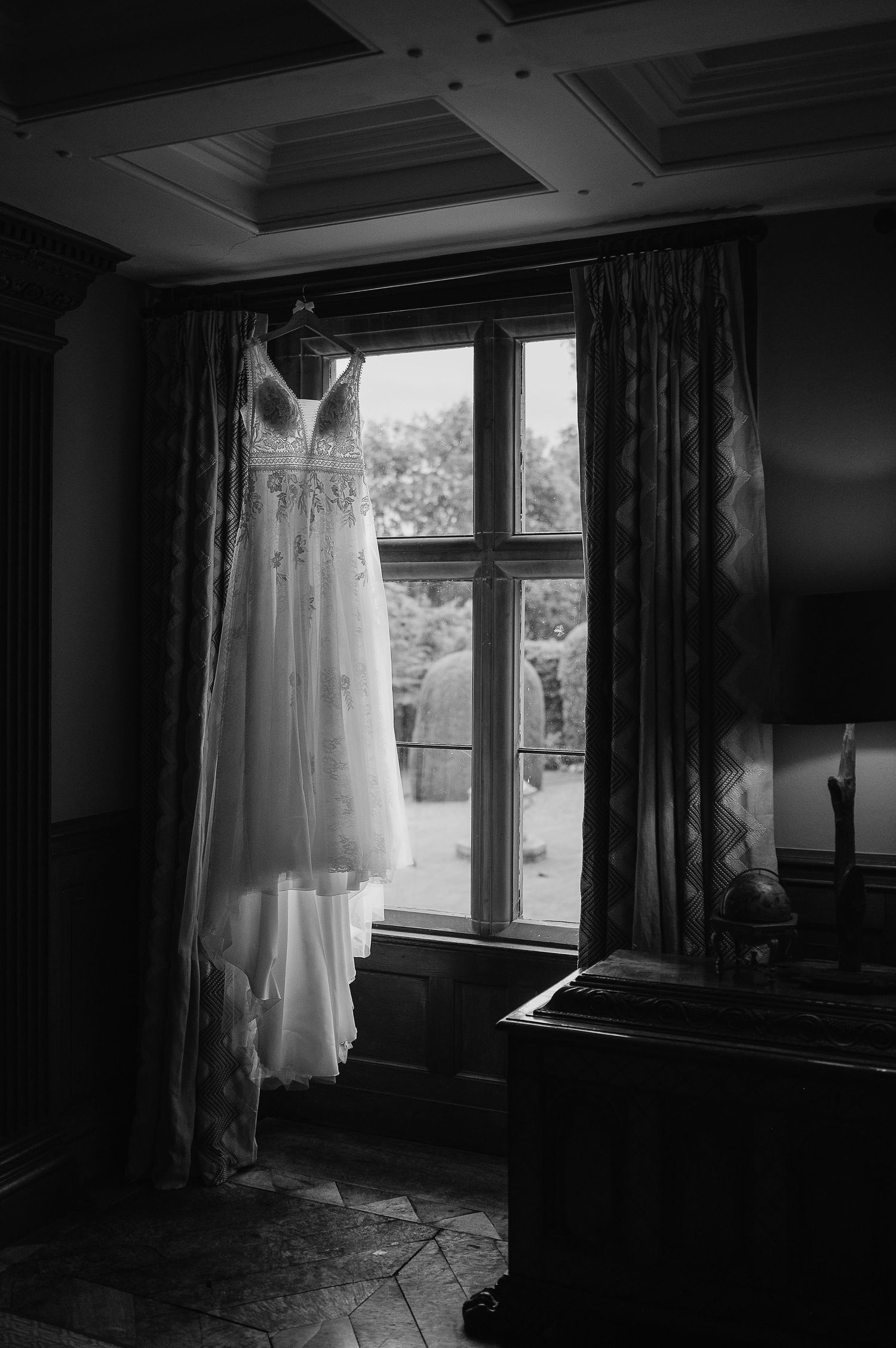 A wedding dress hangs dramatically in front of a window, captured in black and white.