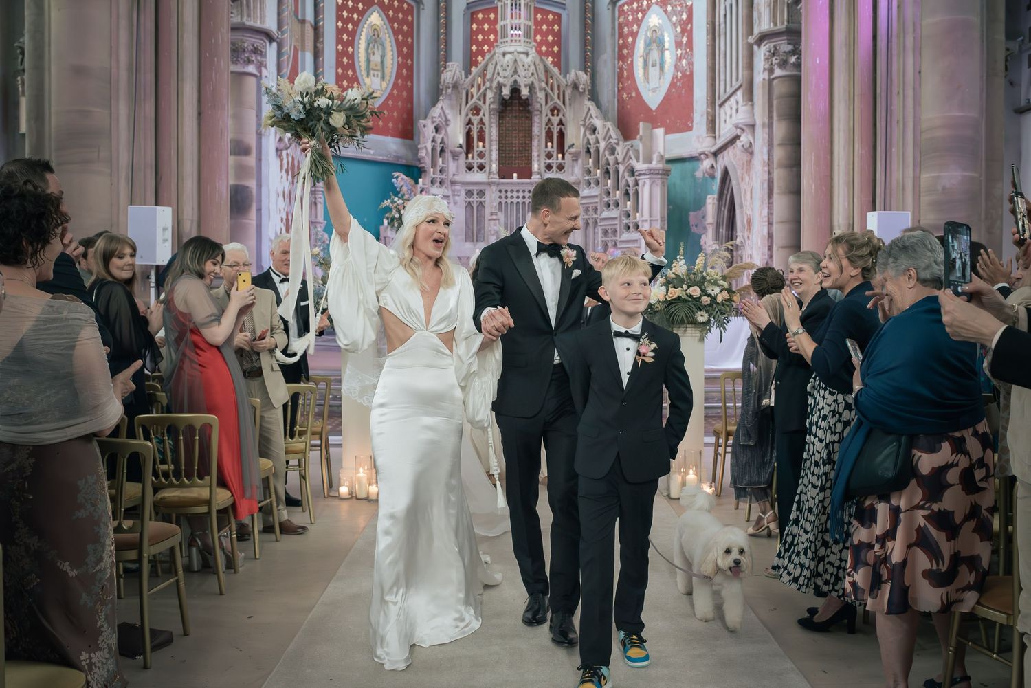 A couple walks down the aisle of an ornate church with red and gold decor while guests look on.