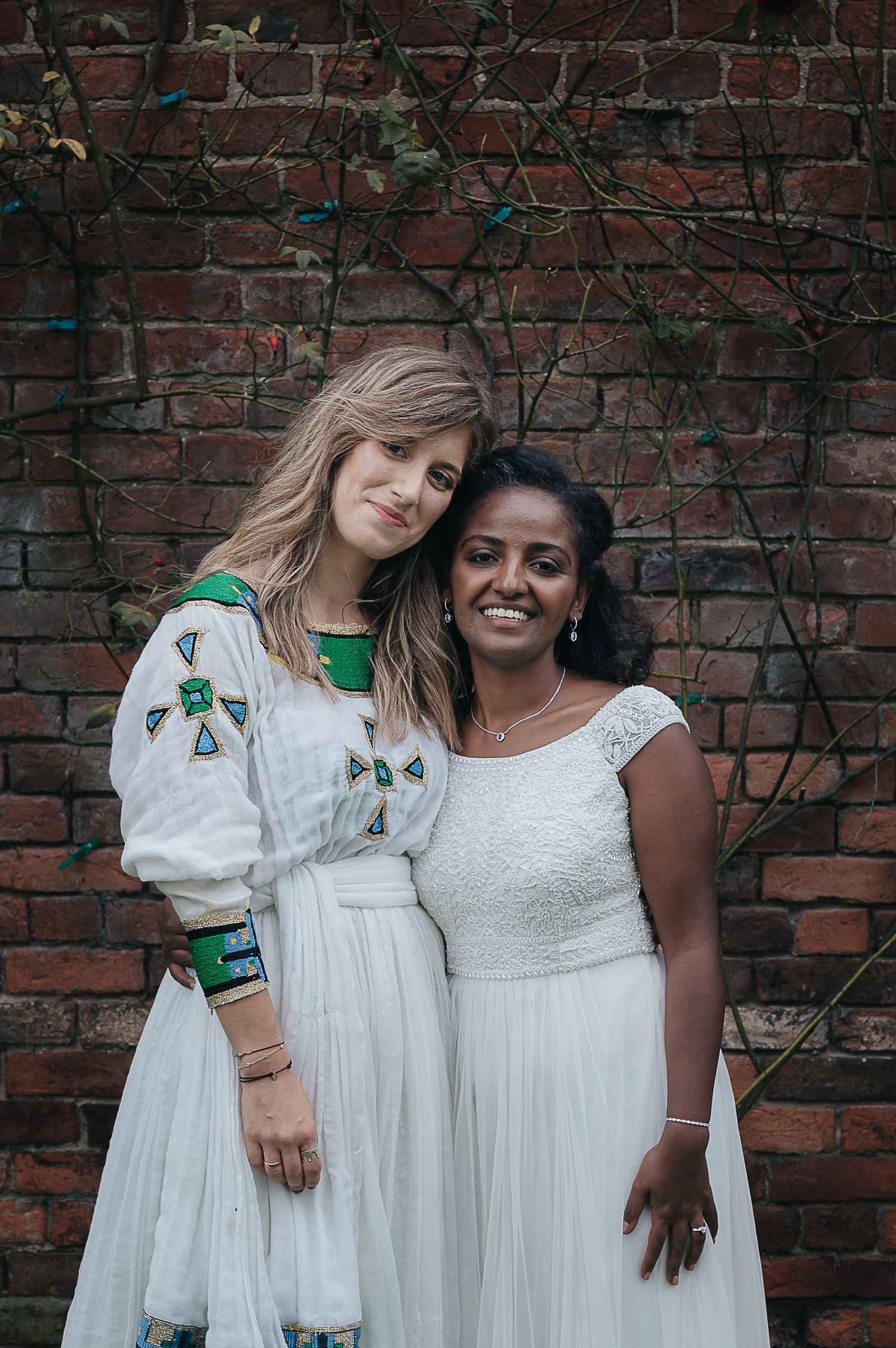 Two people in white dresses pose together against rustic brick wall with climbing vines.