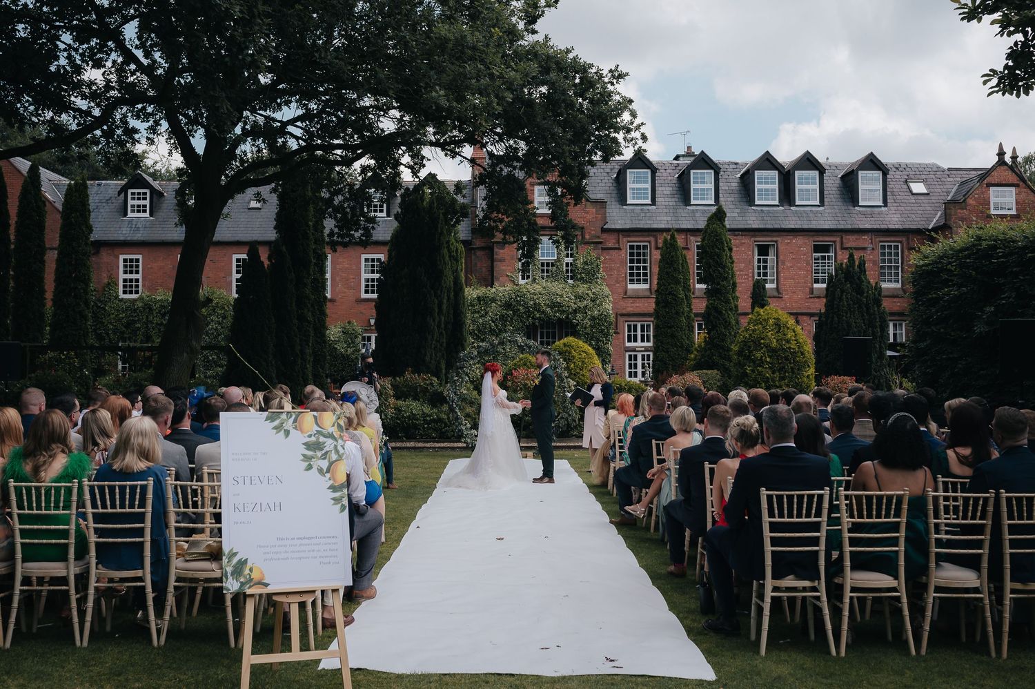 Garden wedding ceremony setup with white aisle runner and gold chairs against backdrop of historic brick building.