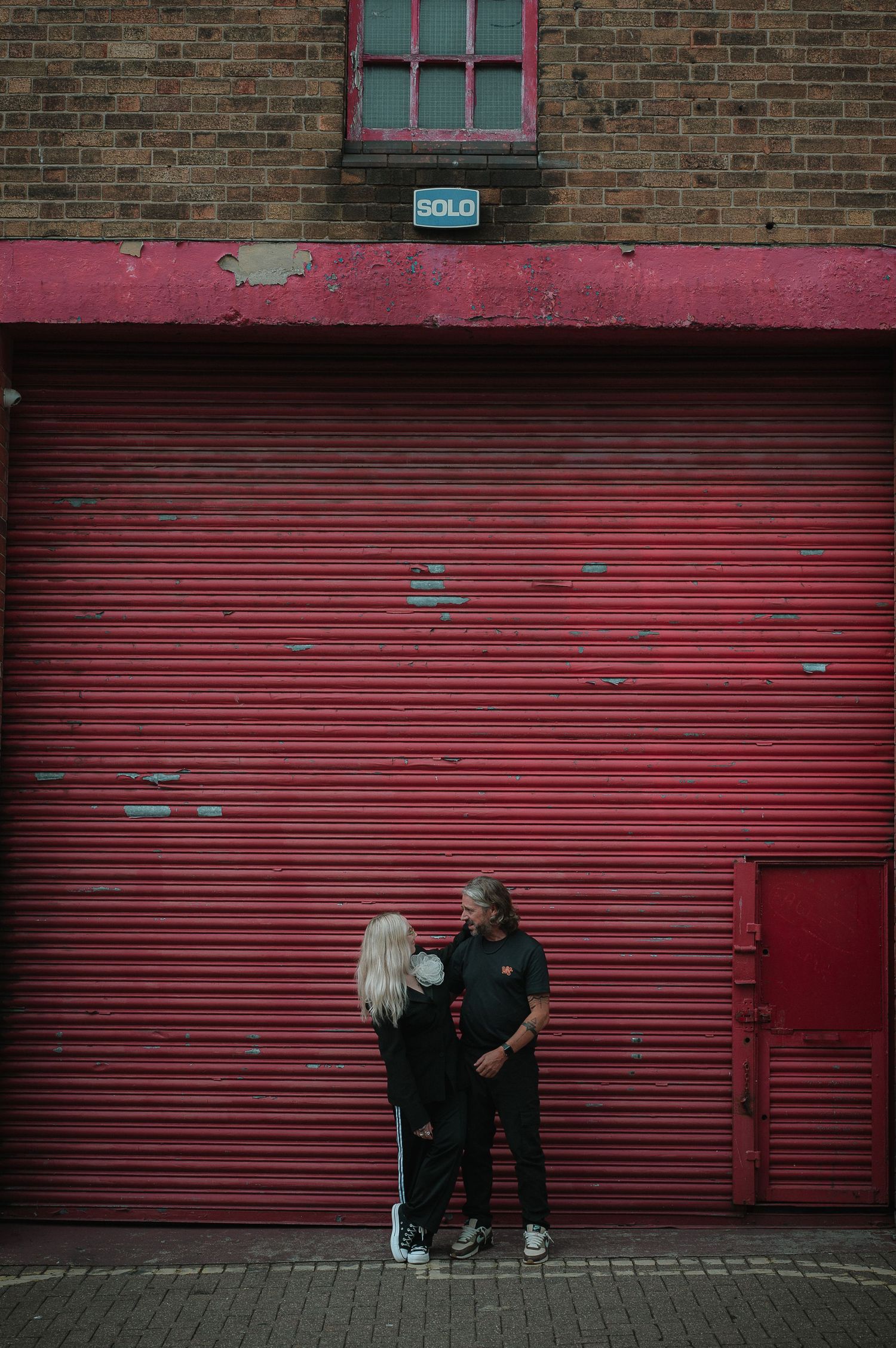 A couple stands intimately against a large red industrial garage door with brick walls in an urban setting.