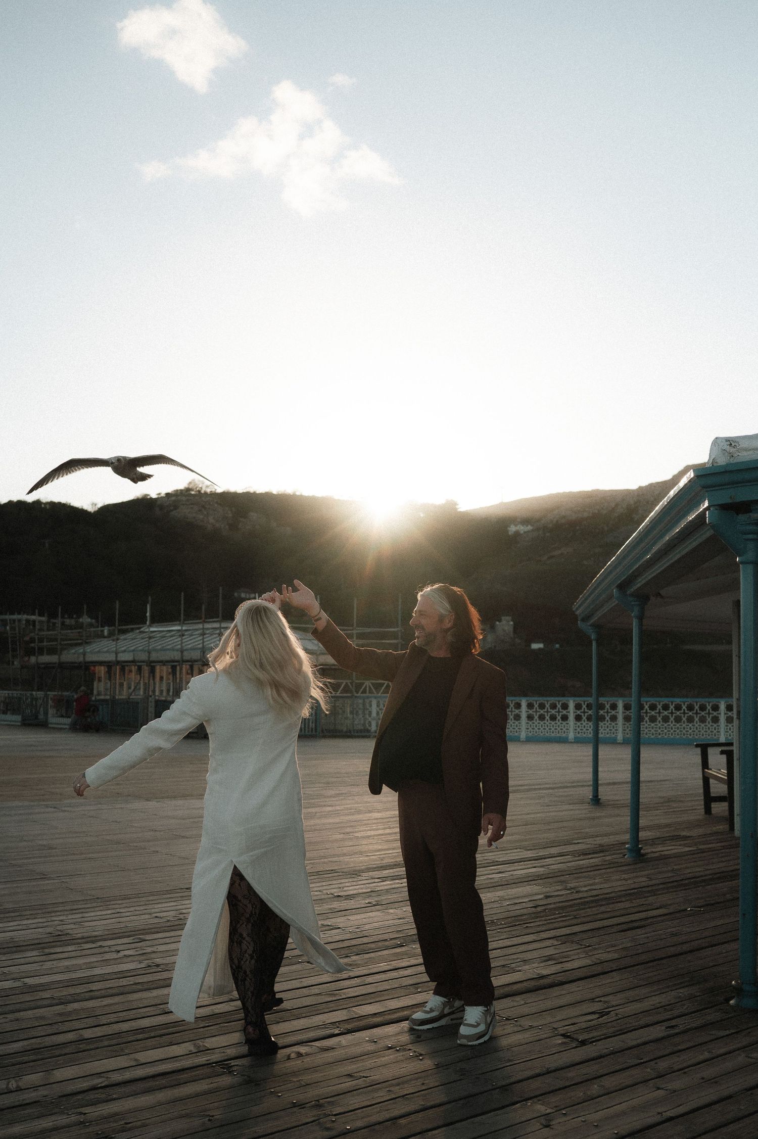 Couple dancing on wooden boardwalk at sunset with seabirds flying overhead.