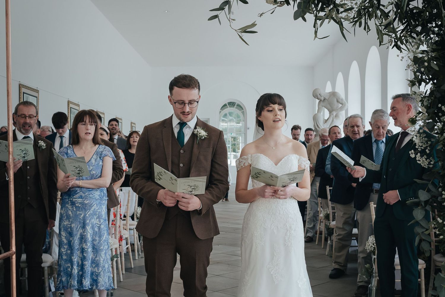 A ceremony taking place in a bright room with arched windows and guests seated in rows.