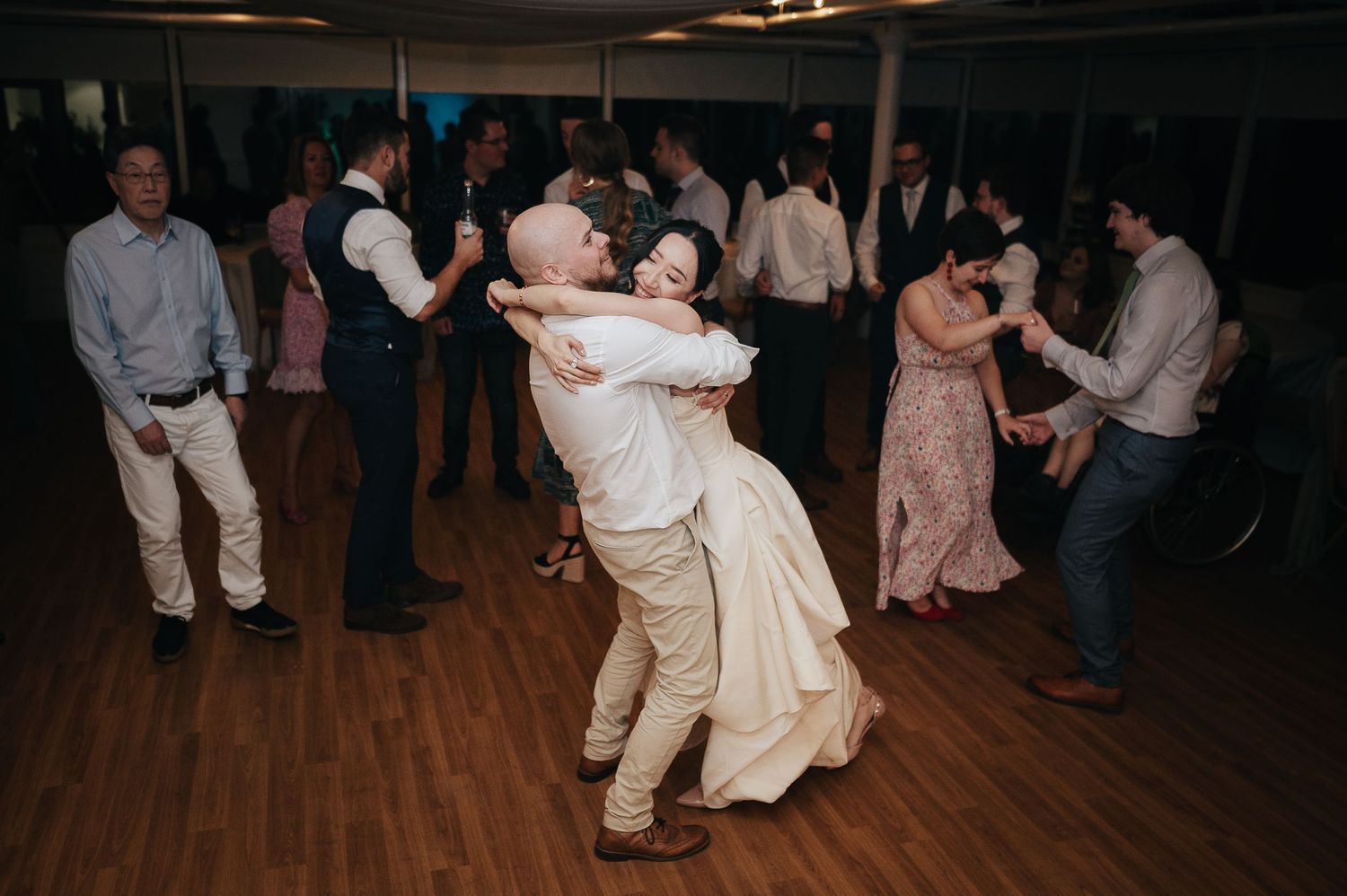 Wedding guests dancing on a wooden floor at an indoor reception venue with dim lighting.