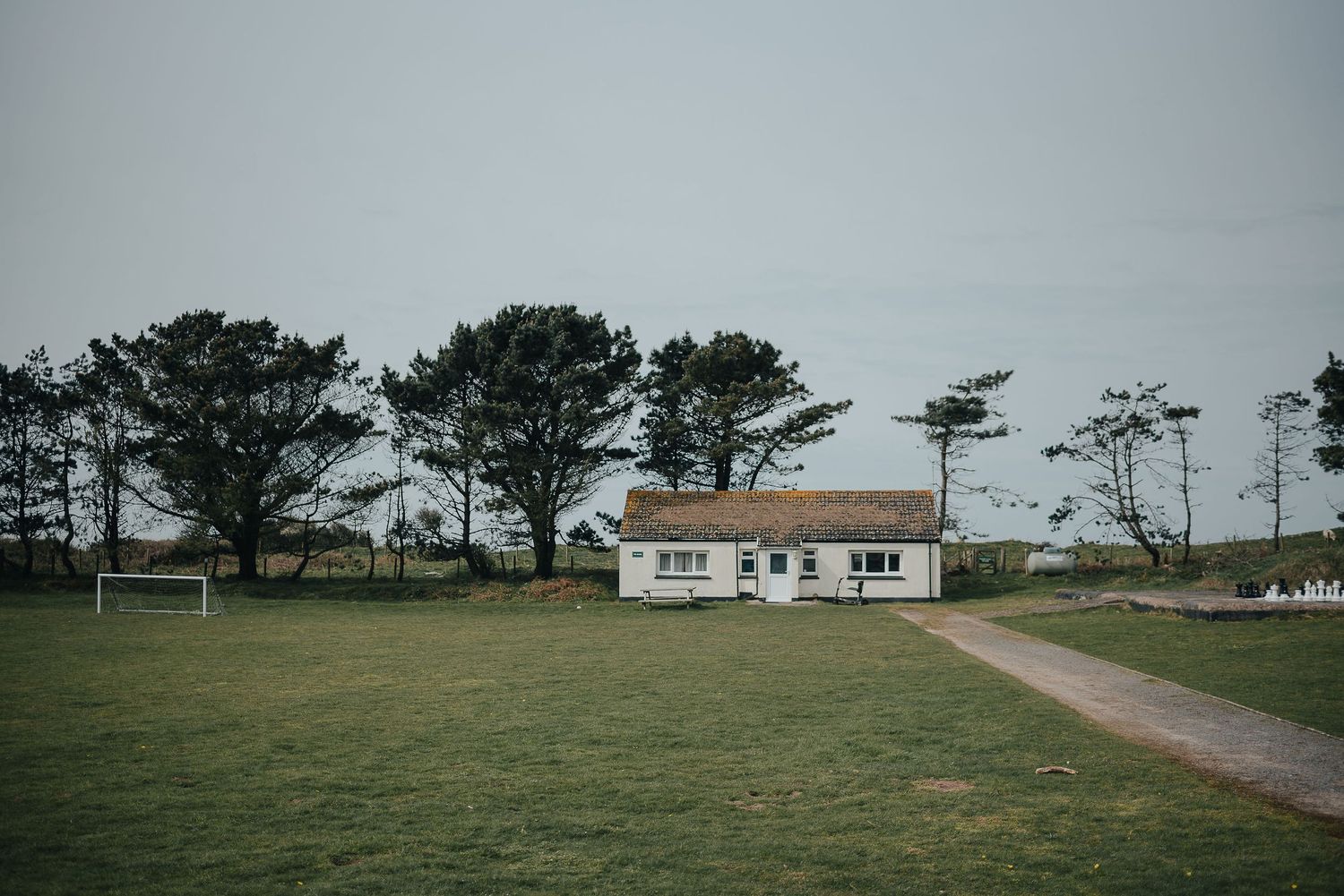 A white single-story building sits isolated on a coastal landscape with windswept pine trees.