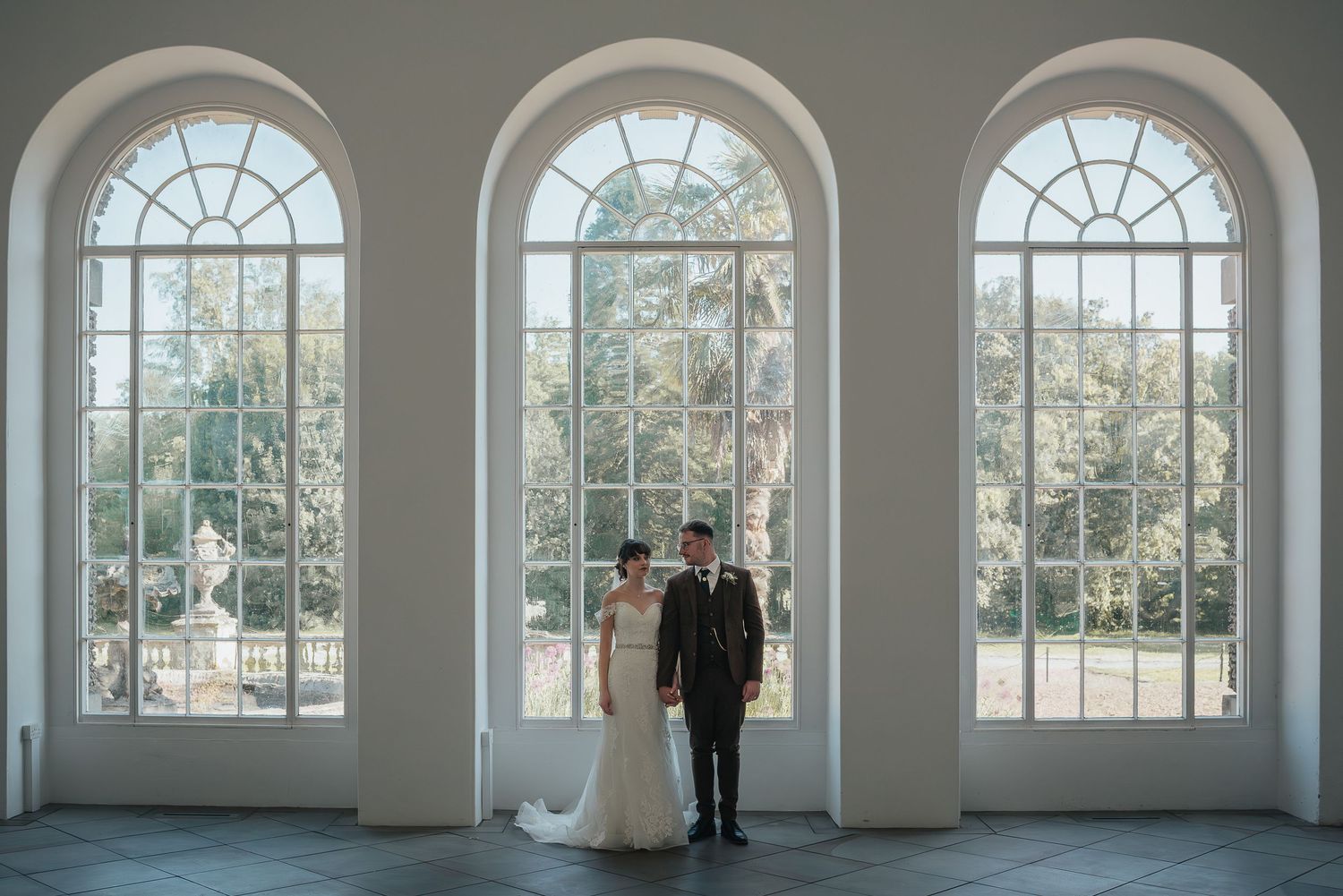 A couple stands silhouetted against three large arched windows in an elegant white room.