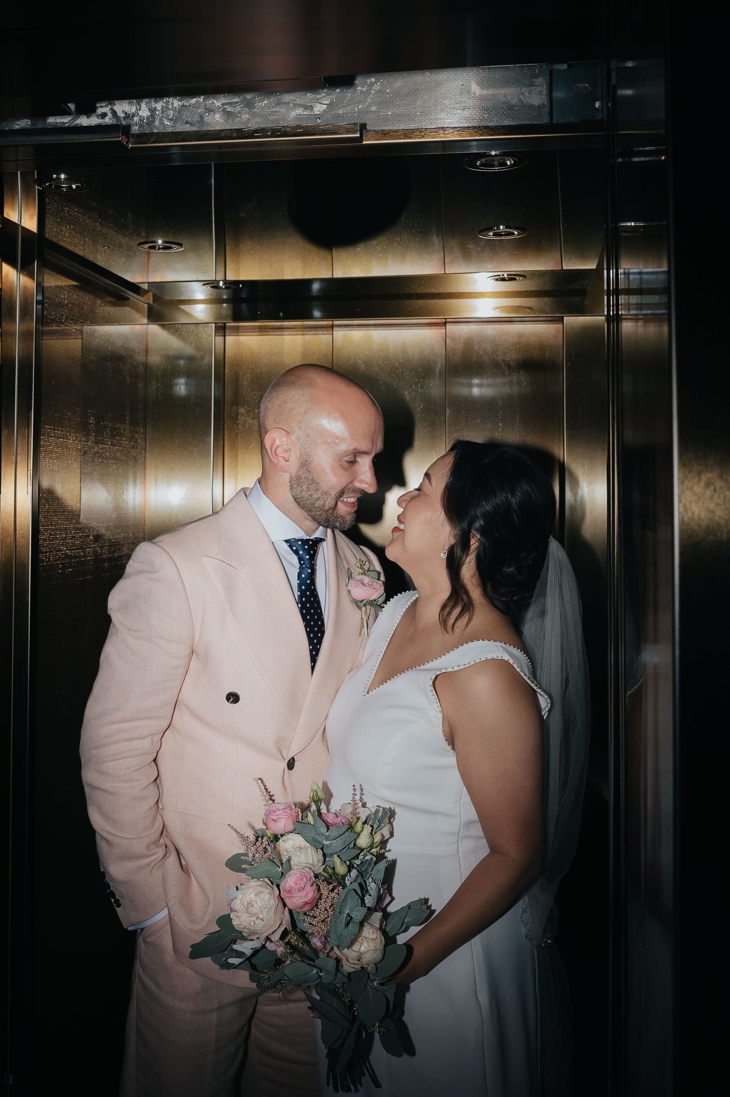 Newlyweds share an intimate moment in a golden-lit elevator holding a floral bouquet.