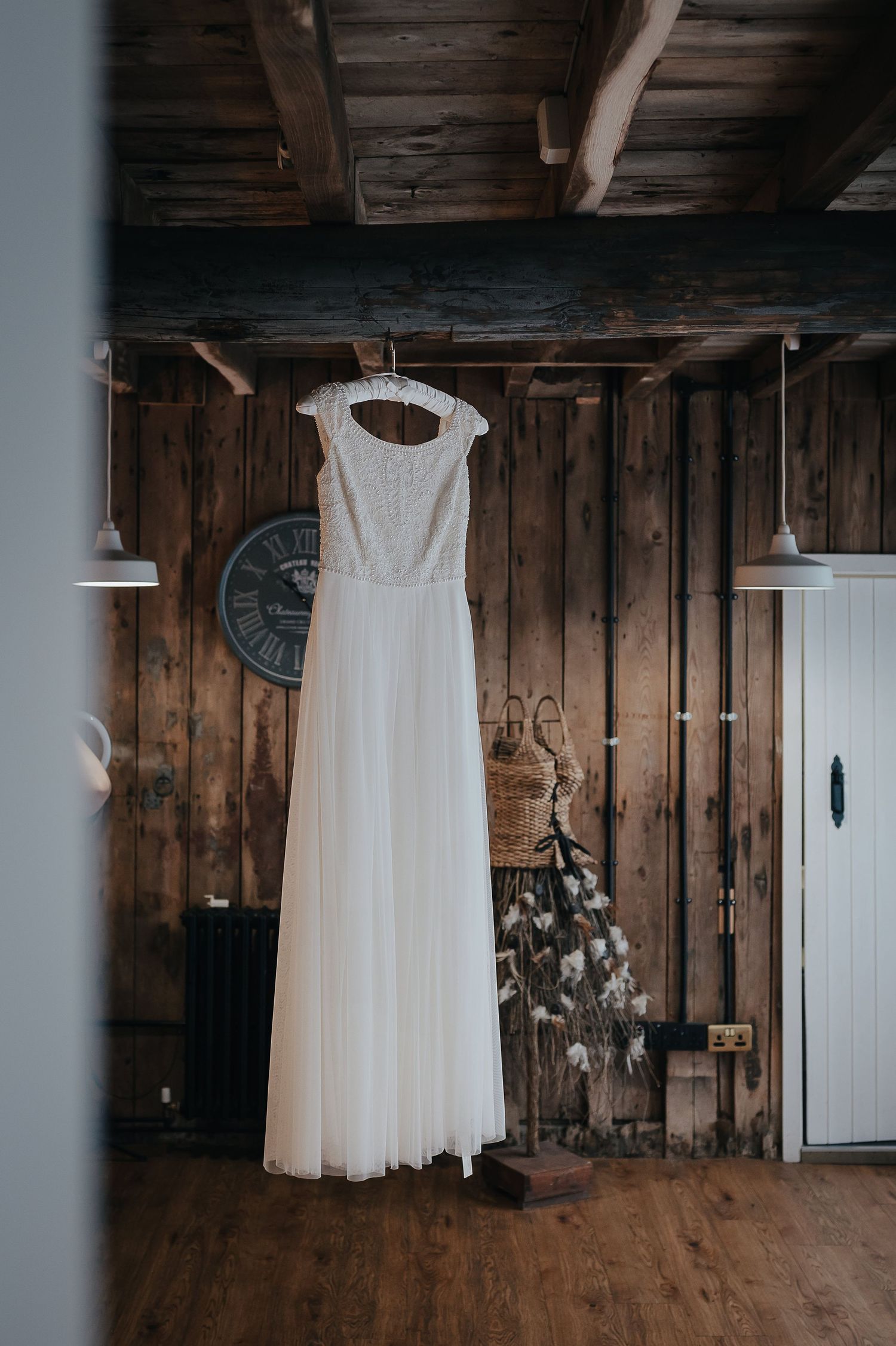 Elegant white wedding dress hangs against rustic wooden wall with vintage clock.