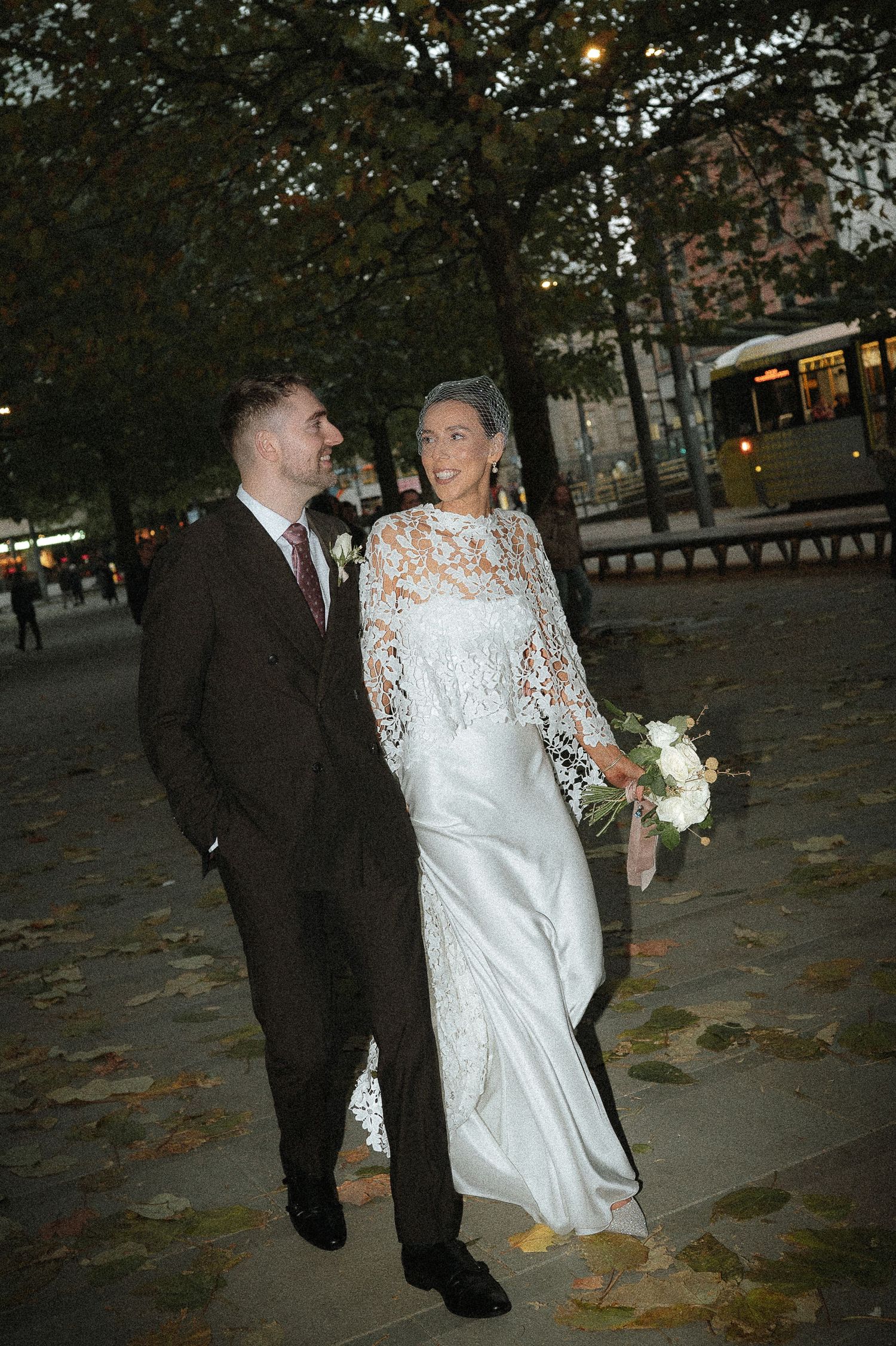 Bride in white lace gown holding bouquet stands with groom in brown suit on leaf-covered path at dusk.